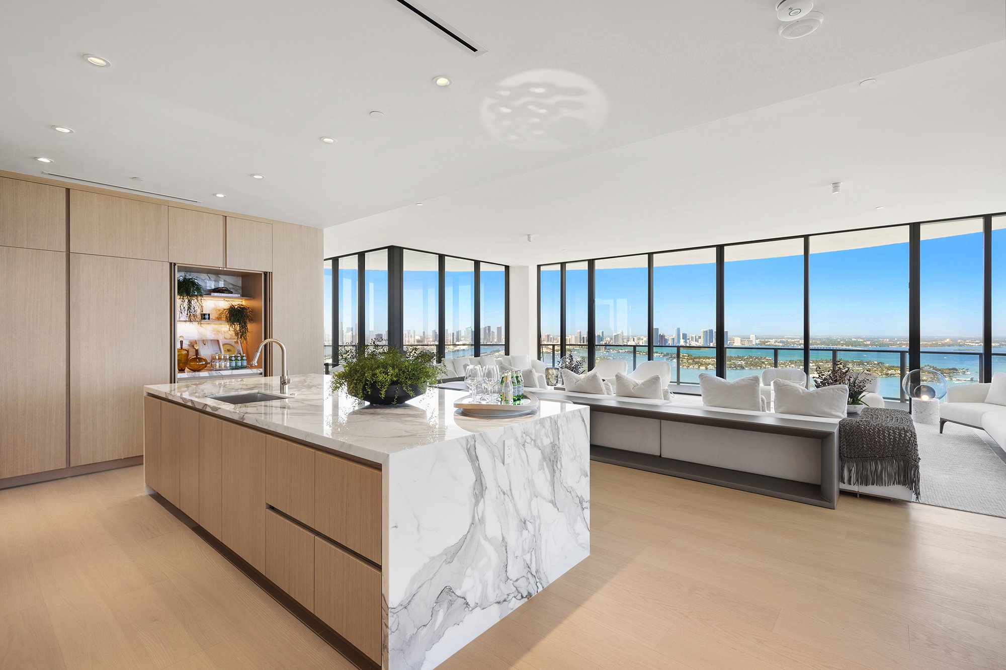 Luxury modern kitchen and living area with marble island, wooden cabinets, and large windows offering city and water views.