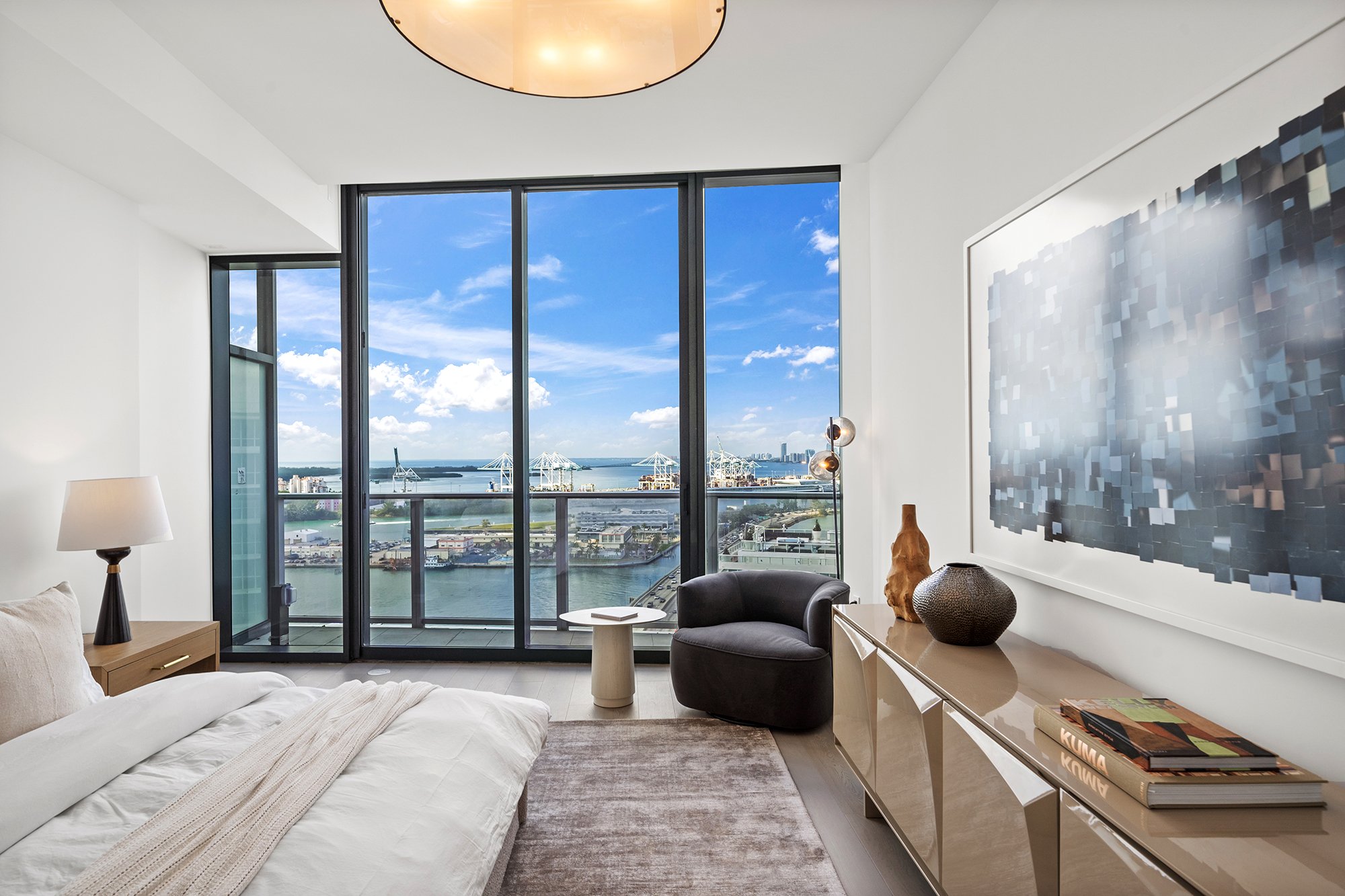 Modern bedroom with large windows, ocean view, bed, side table with lamp, armchair, and abstract wall art.