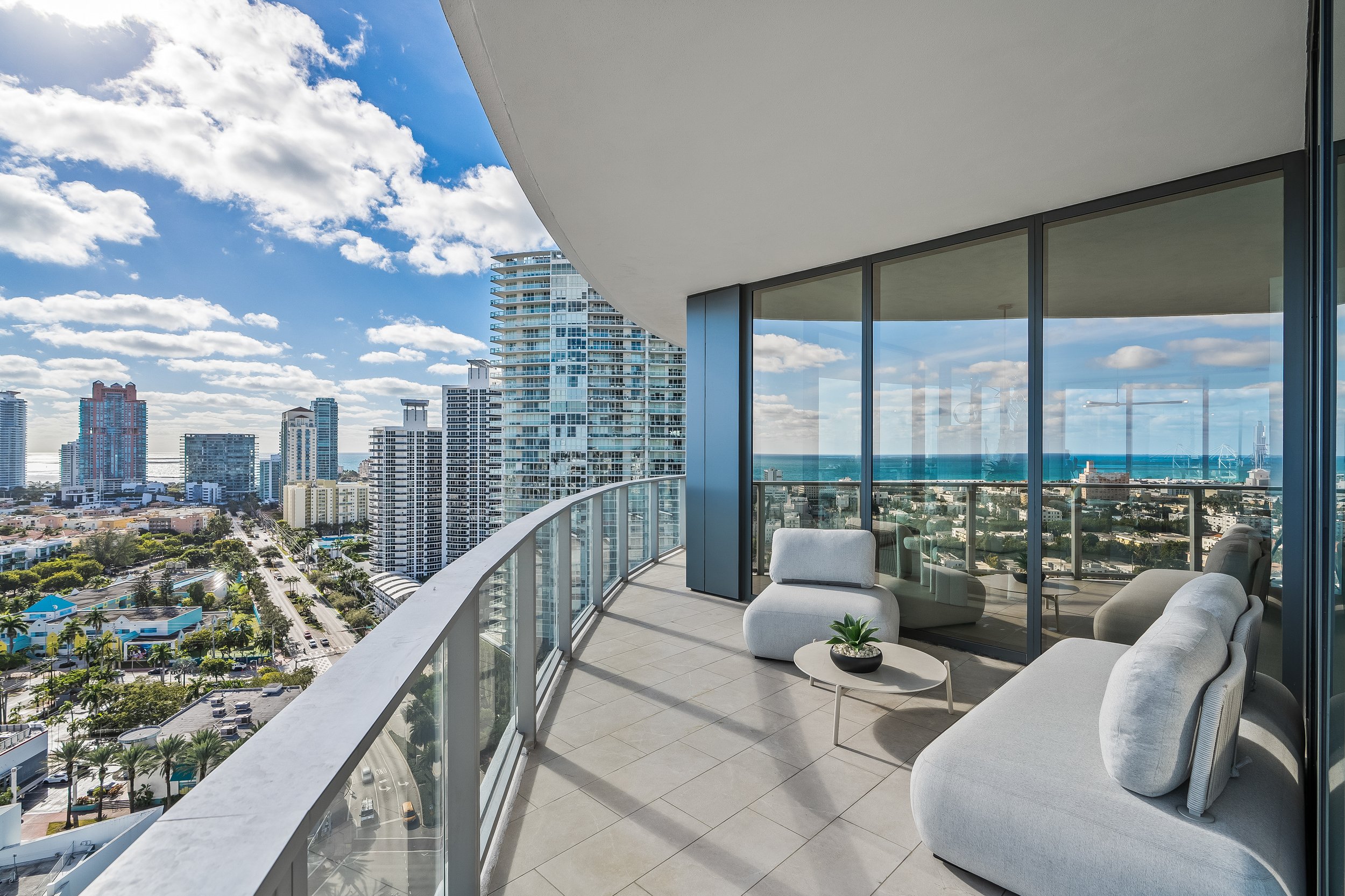 High-rise apartment balcony with city and ocean view, featuring modern outdoor furniture and glass railing.