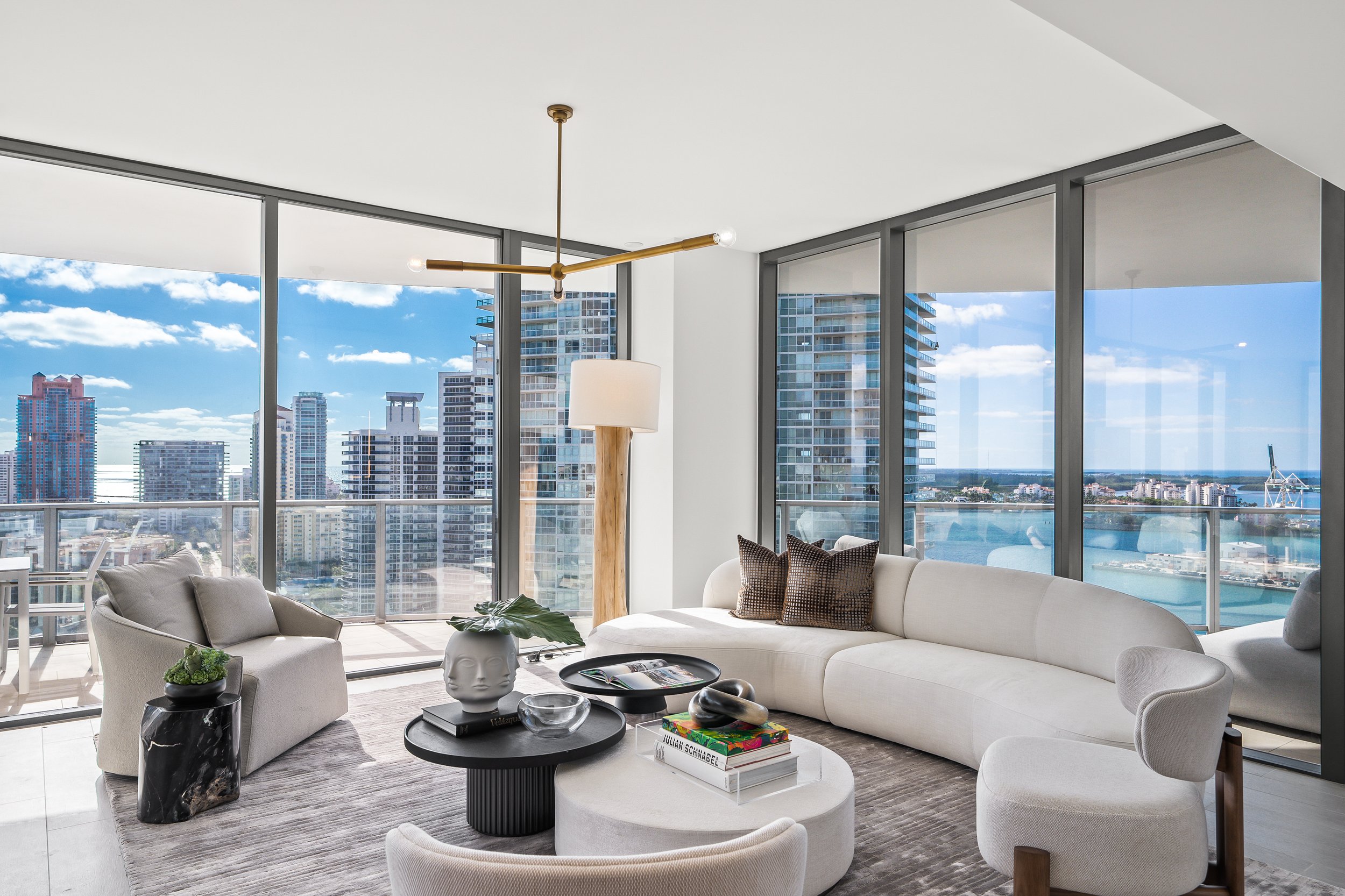 Modern living room with white curved sofa, armchair, and round coffee tables, featuring a large city skyline view through floor-to-ceiling windows.