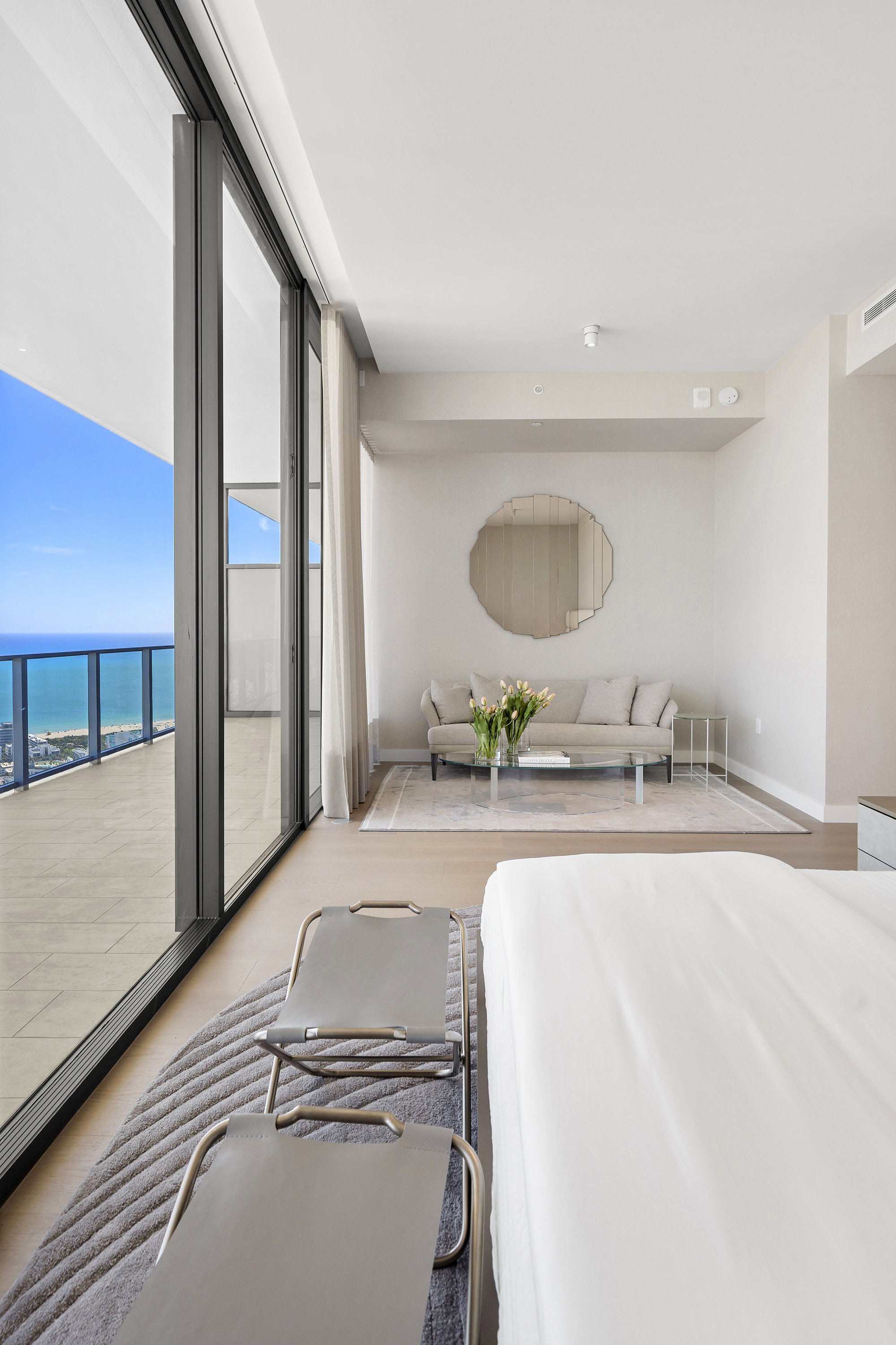 Modern bedroom with large windows overlooking ocean view, featuring a minimalist bed, gray bench, seating area with a couch and glass coffee table, circular mirror on the wall, and neutral decor.