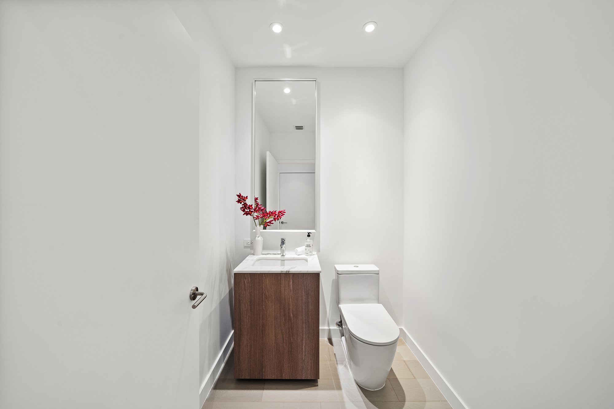 Modern bathroom with toilet, vanity with marble countertop, and a wall mirror, featuring a vase with red flowers and a hand soap dispenser.