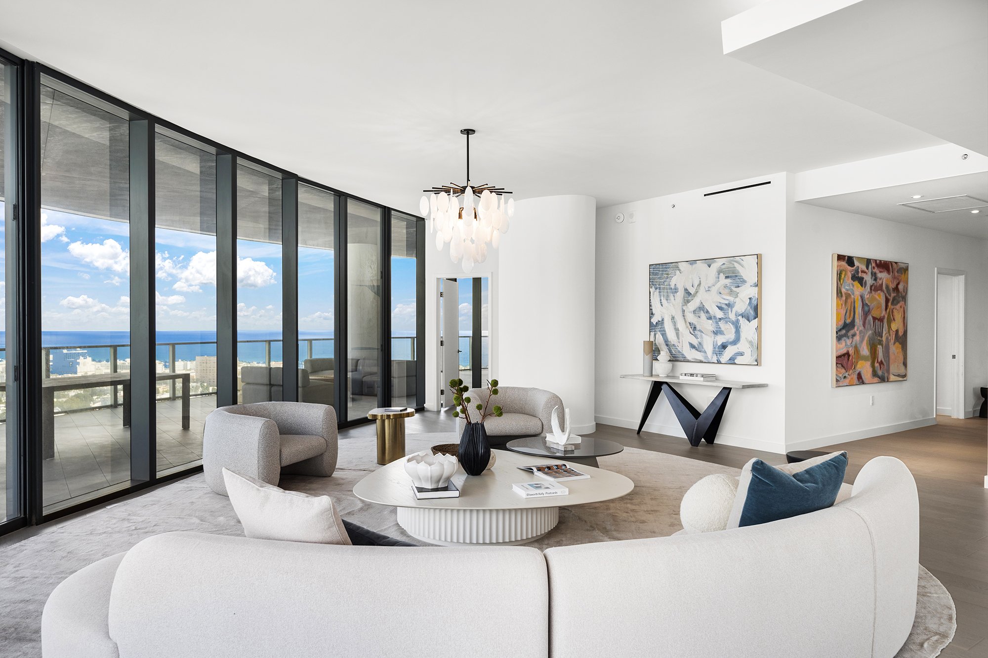 Modern living room with large windows and ocean view, featuring beige curved sofa, round coffee table, contemporary armchairs, abstract art on walls, and a stylish chandelier.