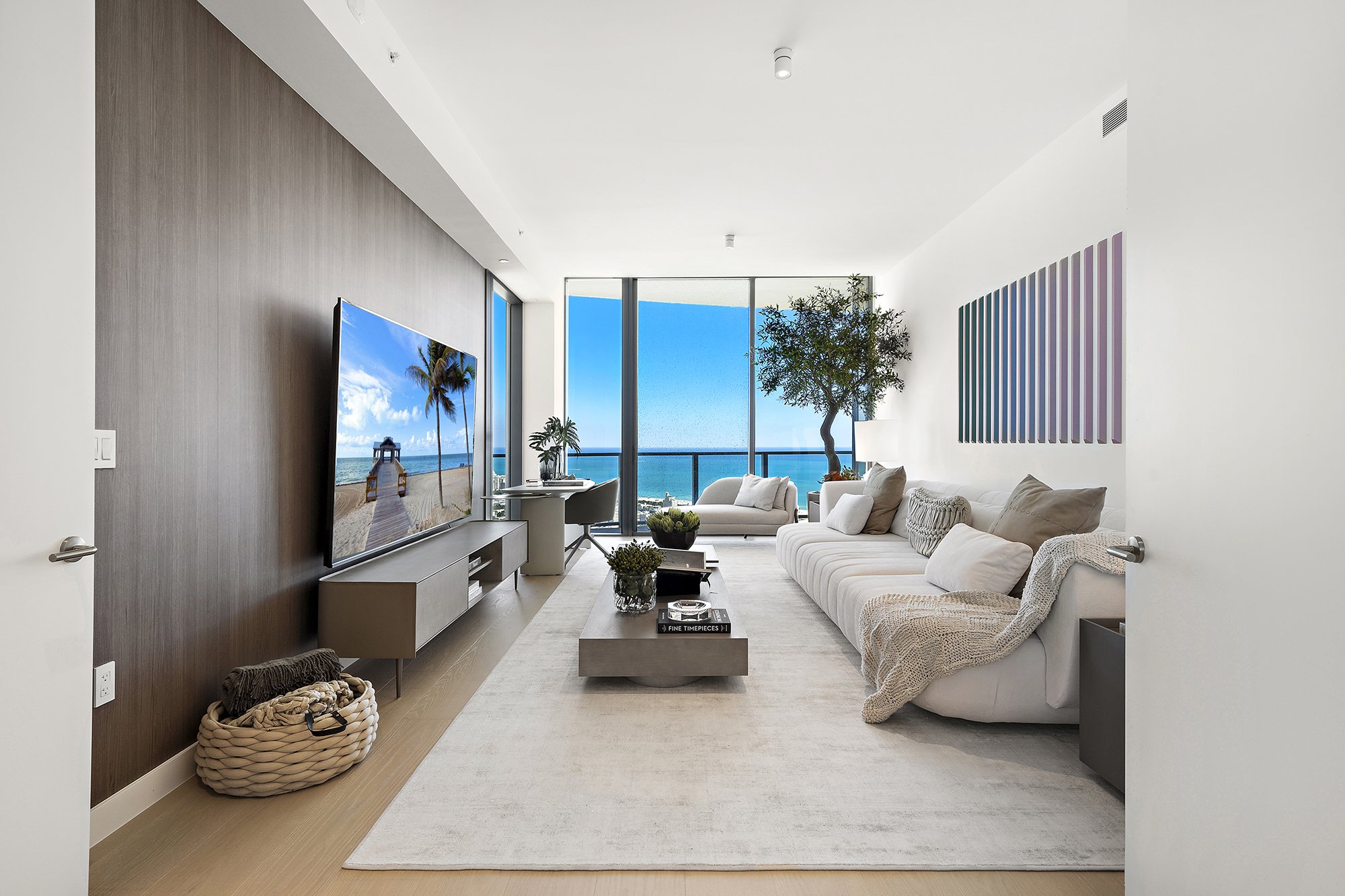 Modern living room with large windows, ocean view, white sectional sofa, TV on a wooden panel wall, indoor plants, coffee table, and decorative basket on the floor.