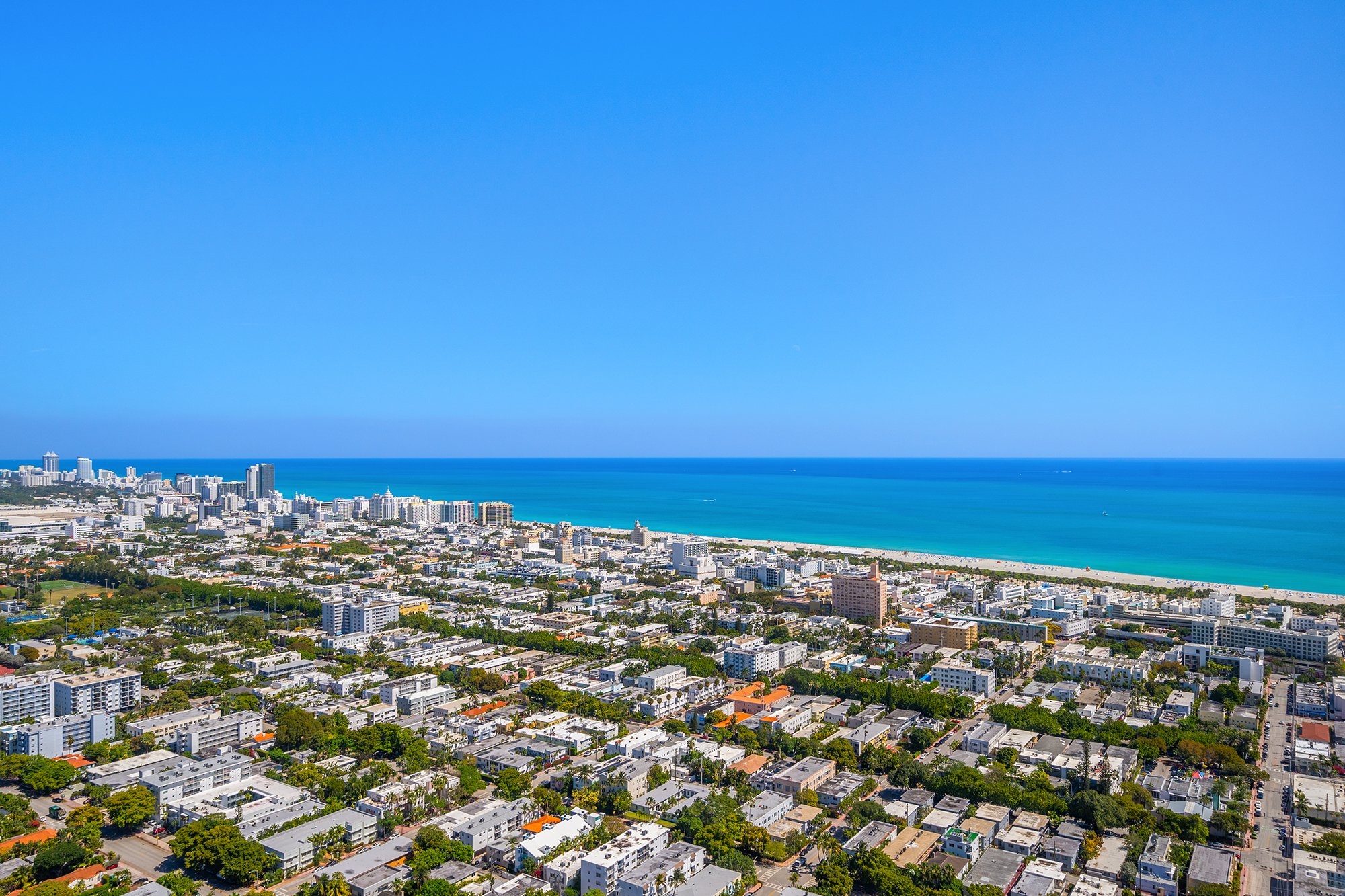 Aerial view of a coastal city with high-rise buildings, residential areas, and a beach along the blue ocean under a clear sky.