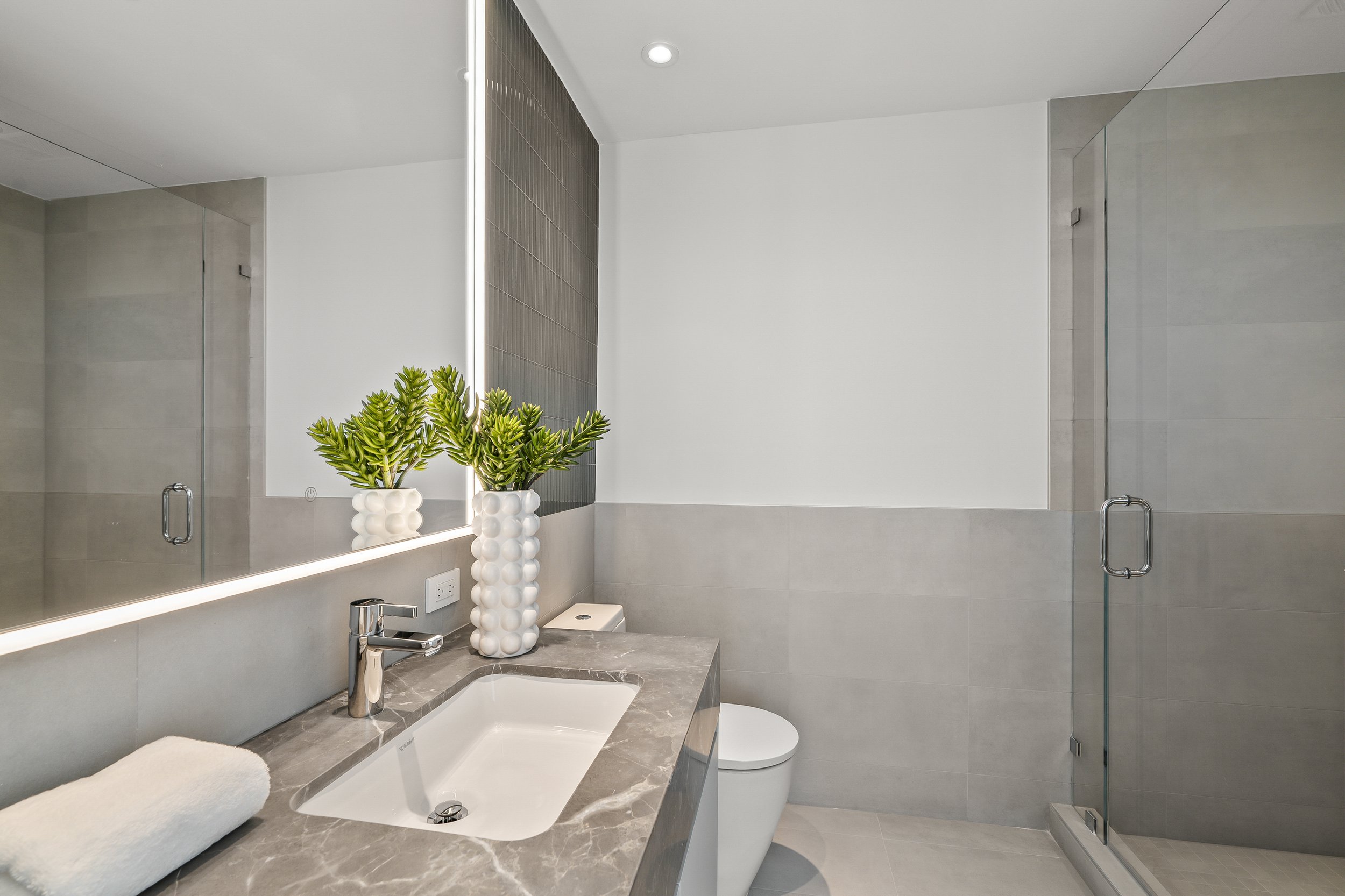 Modern bathroom with marble countertop, white sink, potted plant, illuminated mirror, towel, glass shower, and gray tiles.