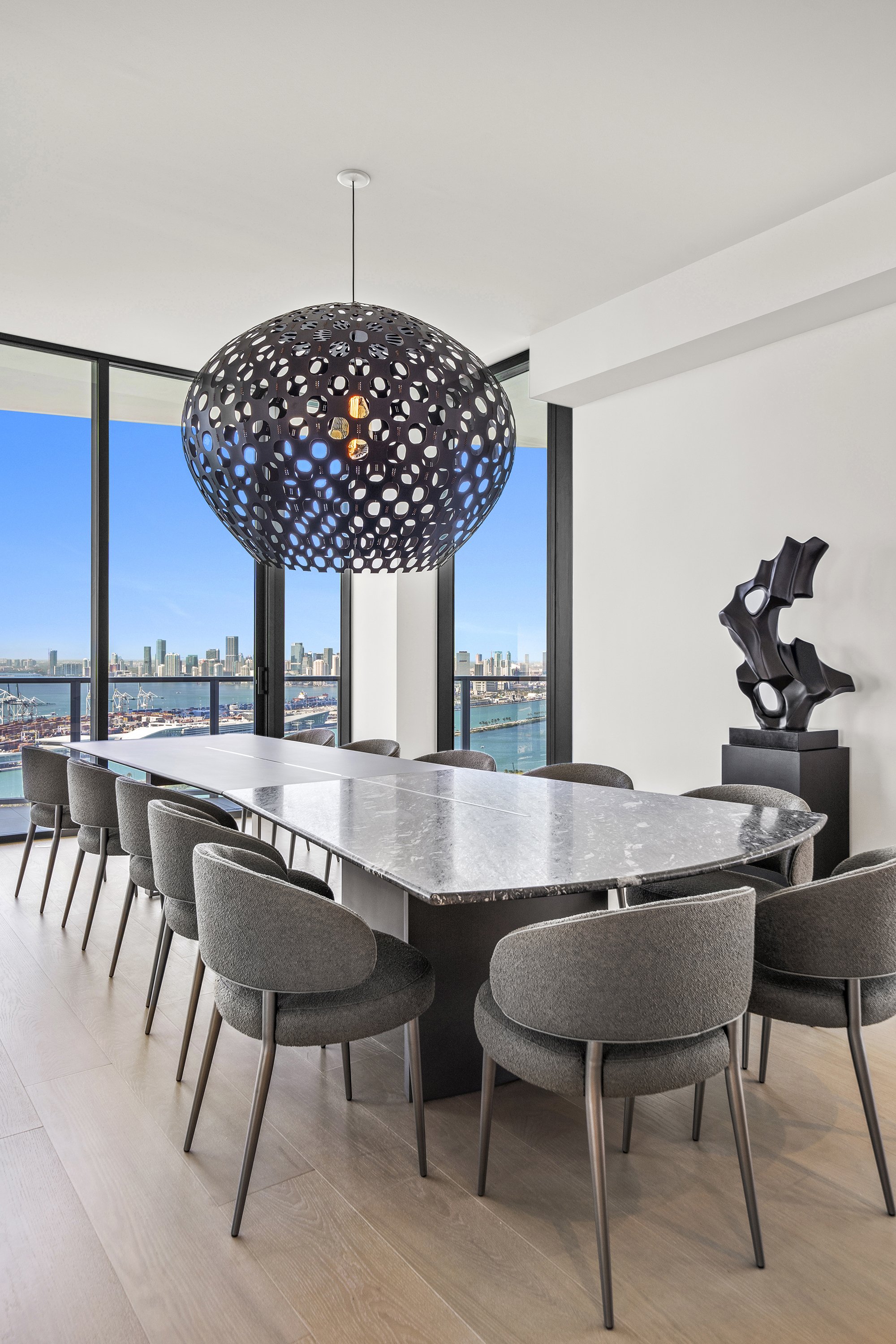 Modern dining room with a long marble table, surrounded by grey chairs with curved backs. Above the table, a large decorative pendant light with a perforated design. Floor-to-ceiling windows offer a cityscape and water view. A black abstract sculptur