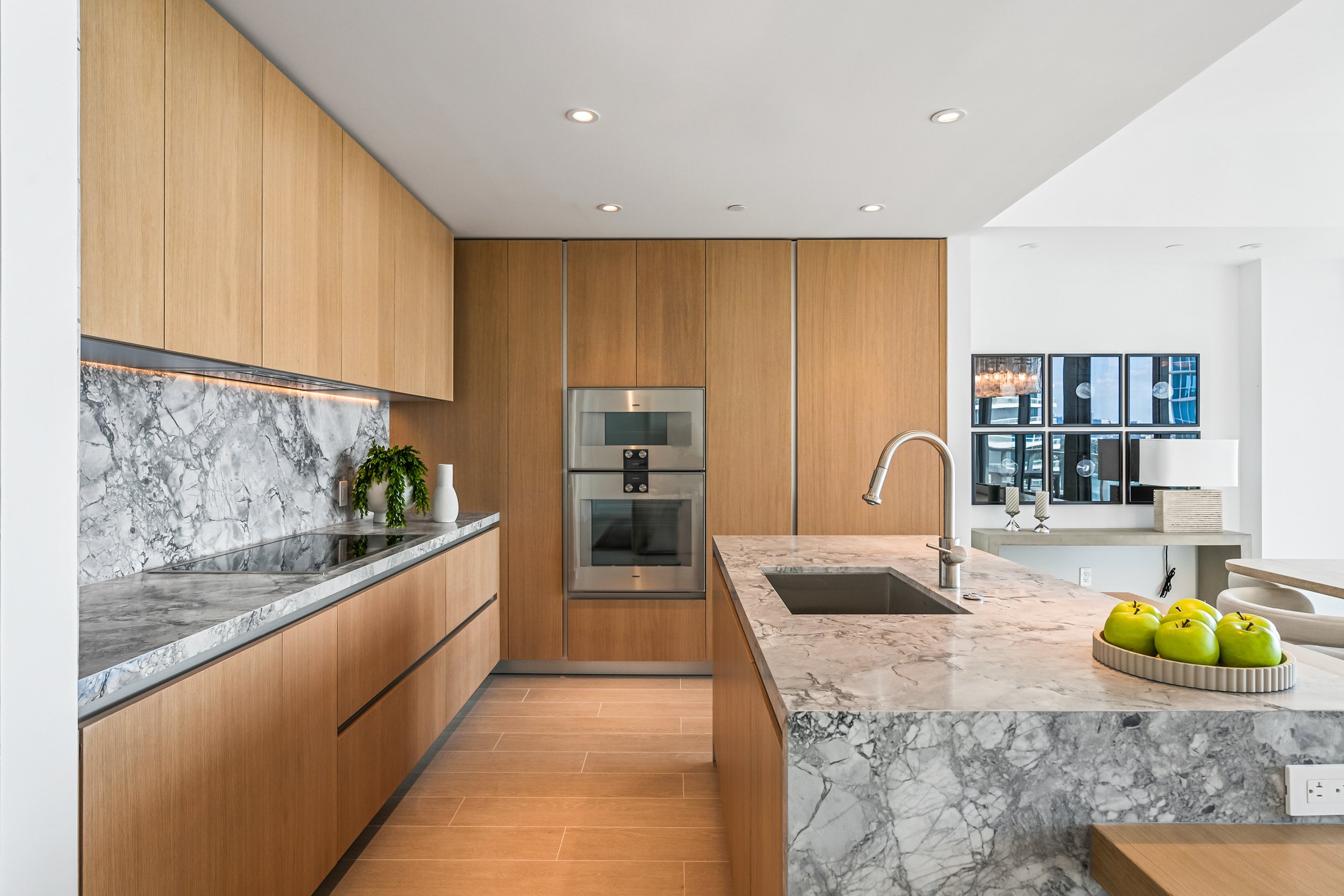 Modern kitchen with marble countertops, wooden cabinetry, stainless steel appliances, and a central island featuring a sink with a faucet. A bowl of green apples sits on the island, and there are decorative plants and accessories throughout.