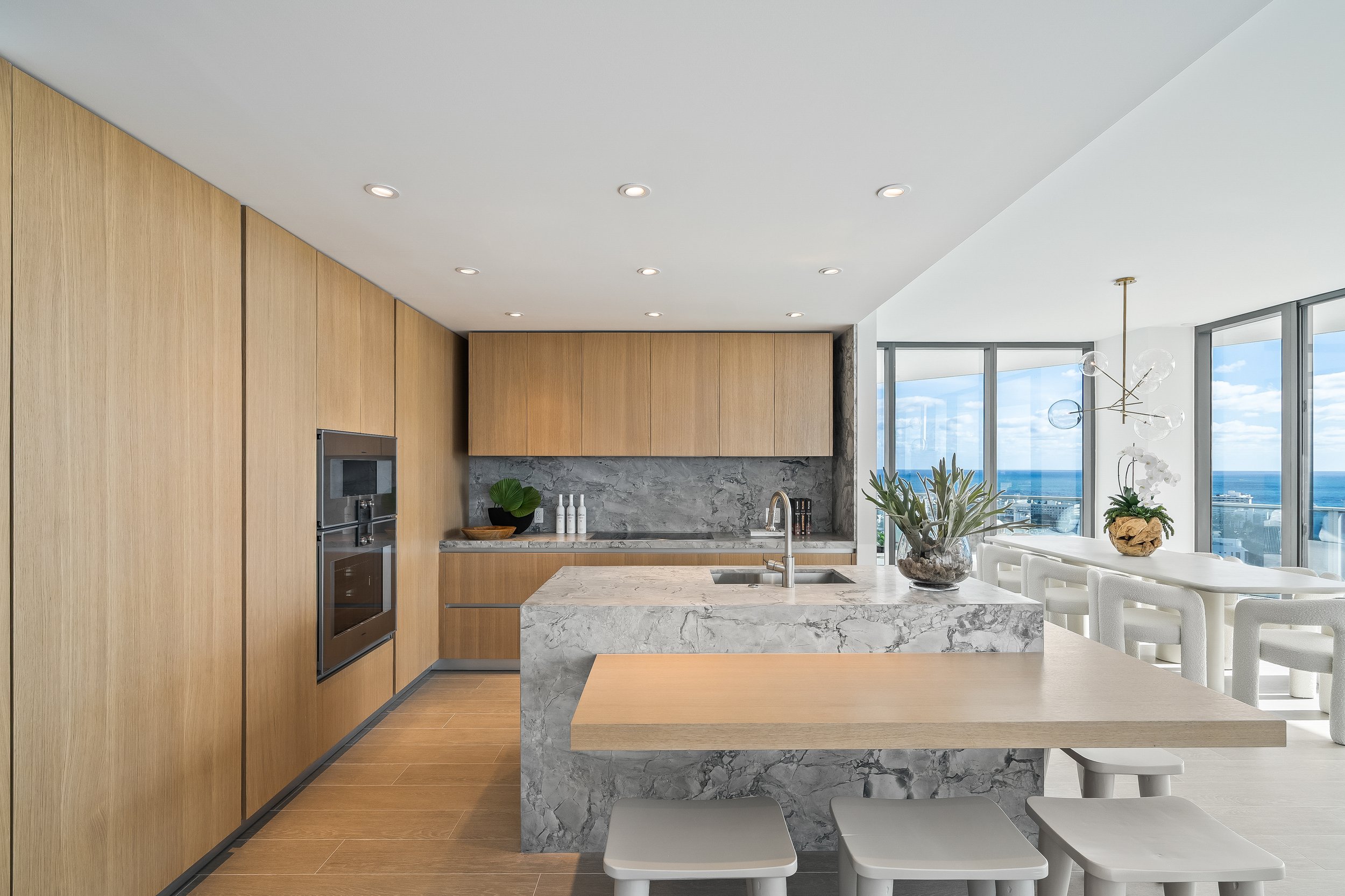 Modern kitchen with wooden cabinets, marble countertops, and a breakfast bar. Includes built-in oven, island sink, bar stools, large windows with ocean view, and contemporary lighting fixture.