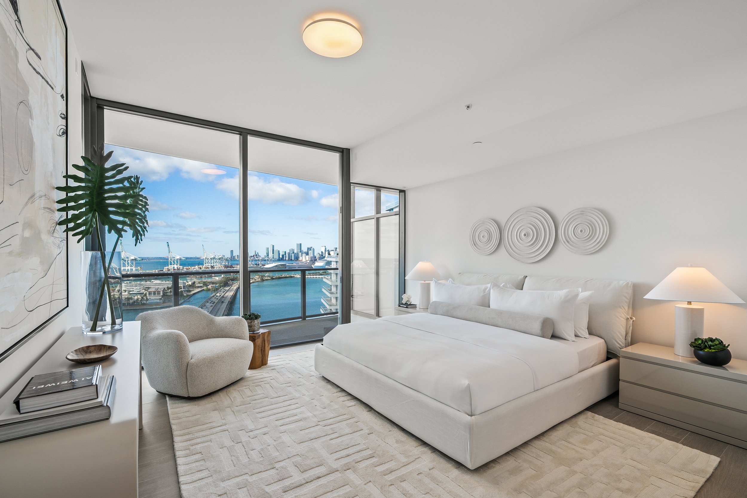 Luxury bedroom with modern decor, large windows offering city and water views, and a plush chair in a corner.