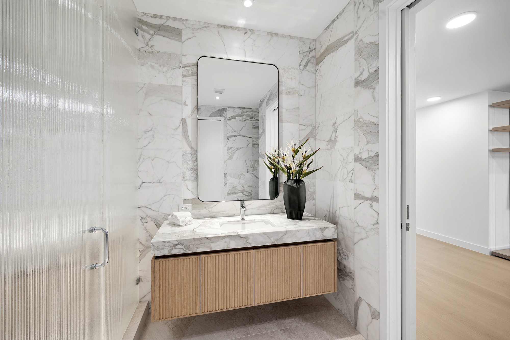 Modern bathroom with marble walls, a large mirror, a marble countertop with a sink, a black vase with flowers, and wood paneling.