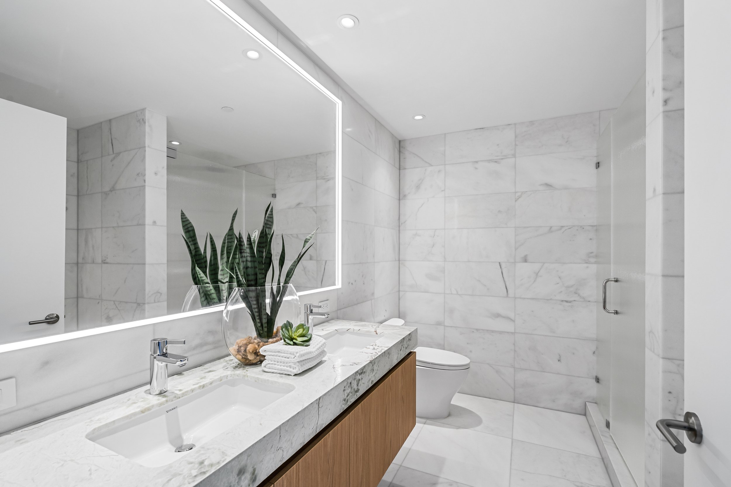Modern bathroom with white marble tiles, a large mirror with integrated lighting, twin sinks on a marble countertop, plants, and a glass shower door.