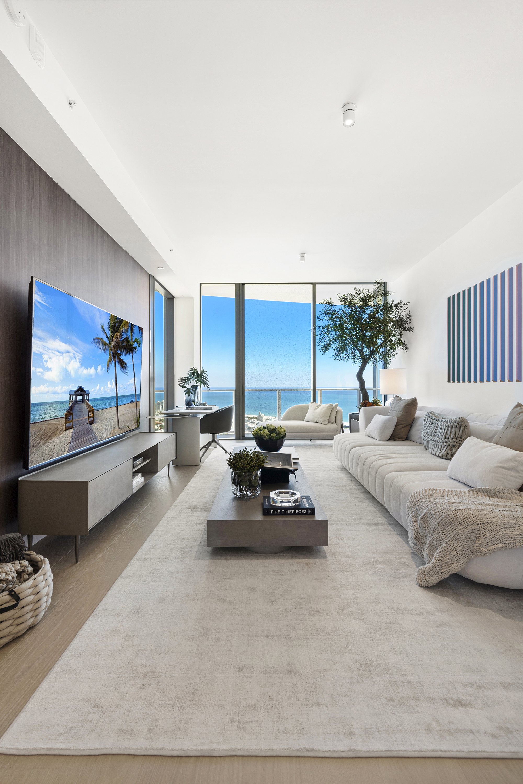 Modern living room with large windows overlooking ocean, featuring a beige sectional sofa, coffee table, flat-screen TV, and decorative plants.