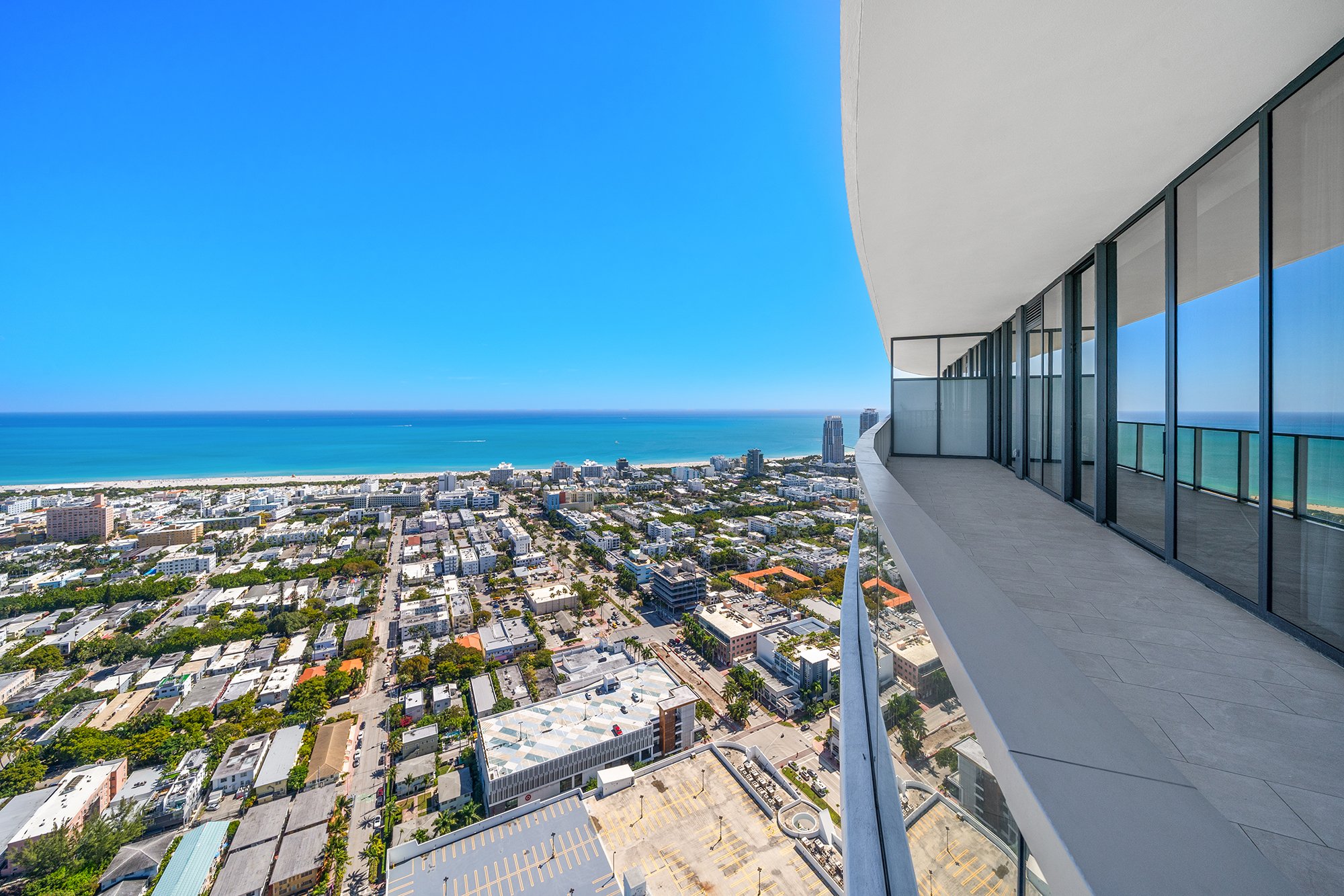 Aerial view from a high-rise building balcony overlooking a cityscape with a blue ocean and sky in the background. The balcony features glass railings and tiled flooring.