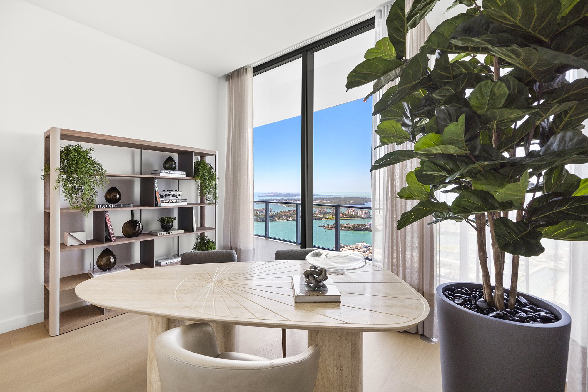 Modern interior with a wooden table, chairs, bookcase with decorative items, large plant, and a floor-to-ceiling window with ocean view.