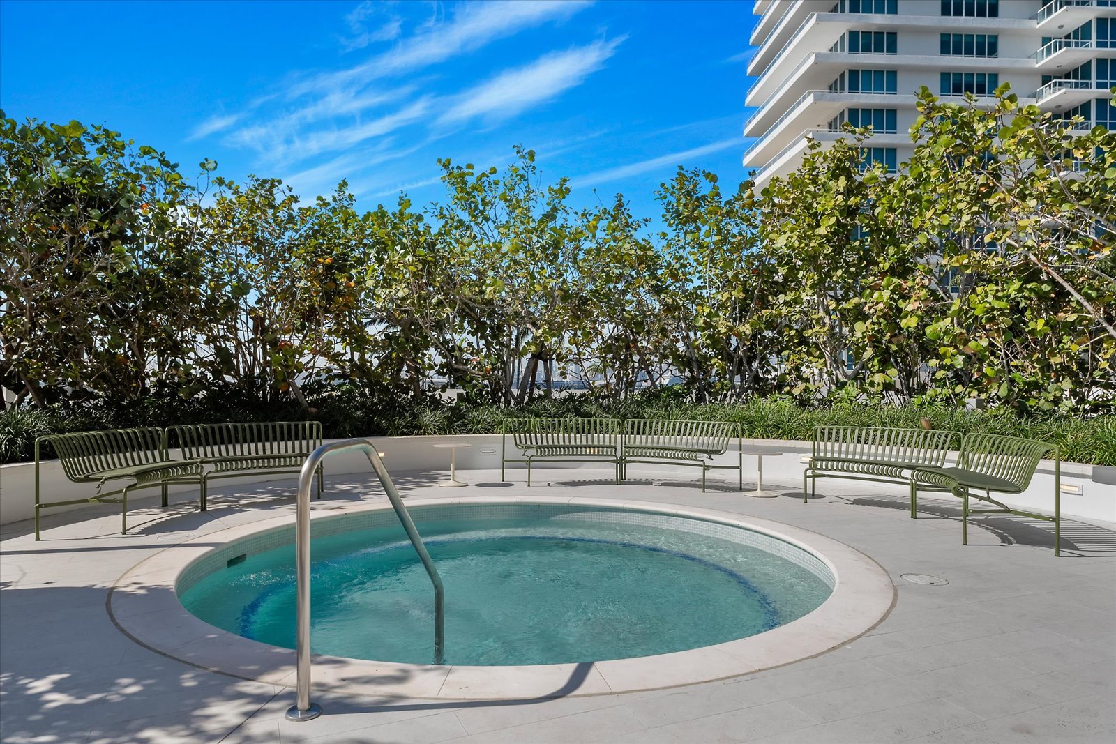 Outdoor jacuzzi with surrounding green benches and lush greenery, set against a backdrop of a modern building and clear blue sky.