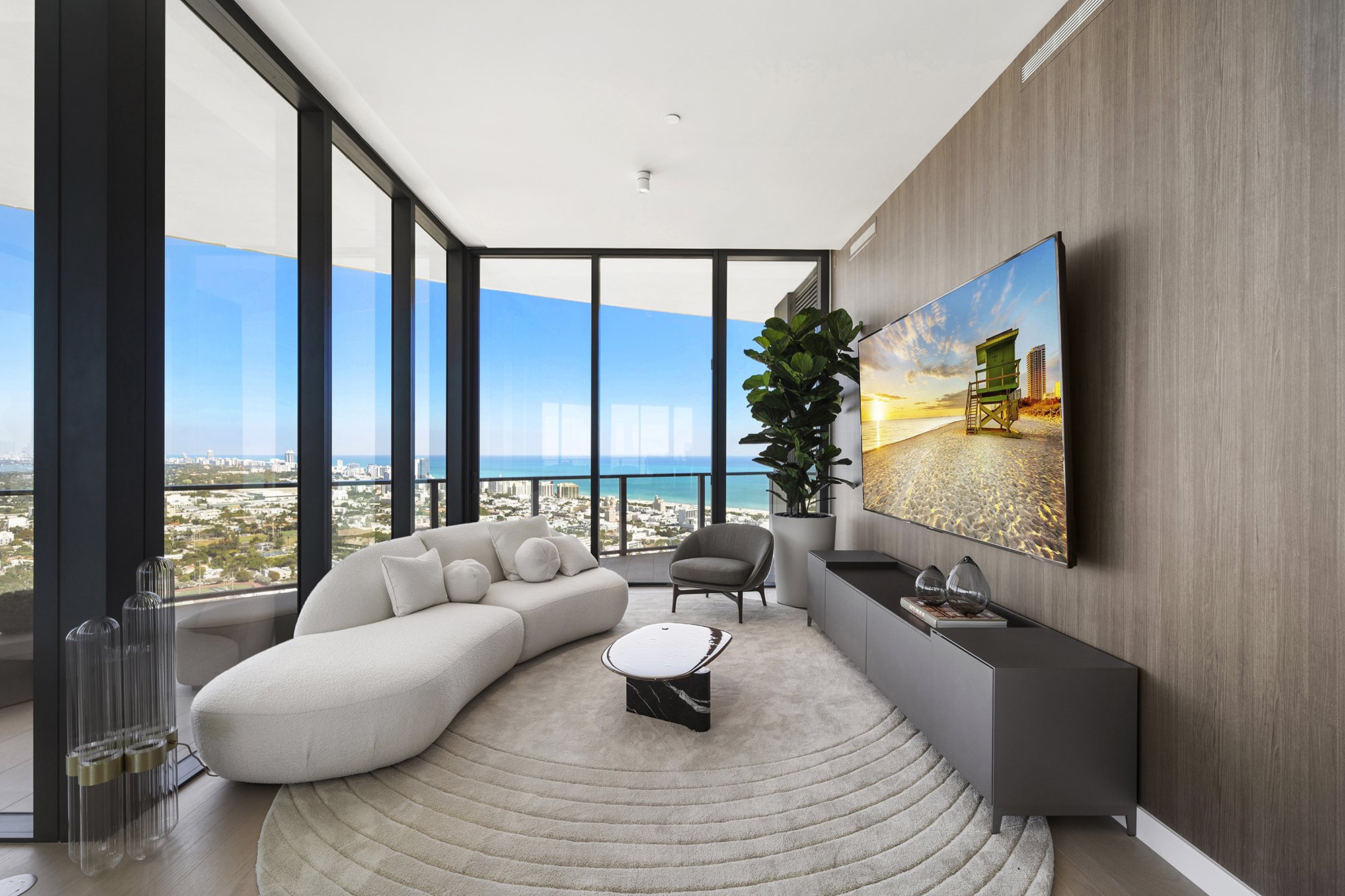 Modern living room with large windows, white sofa, armchair, round coffee table, plant, wall-mounted TV, city and ocean view.