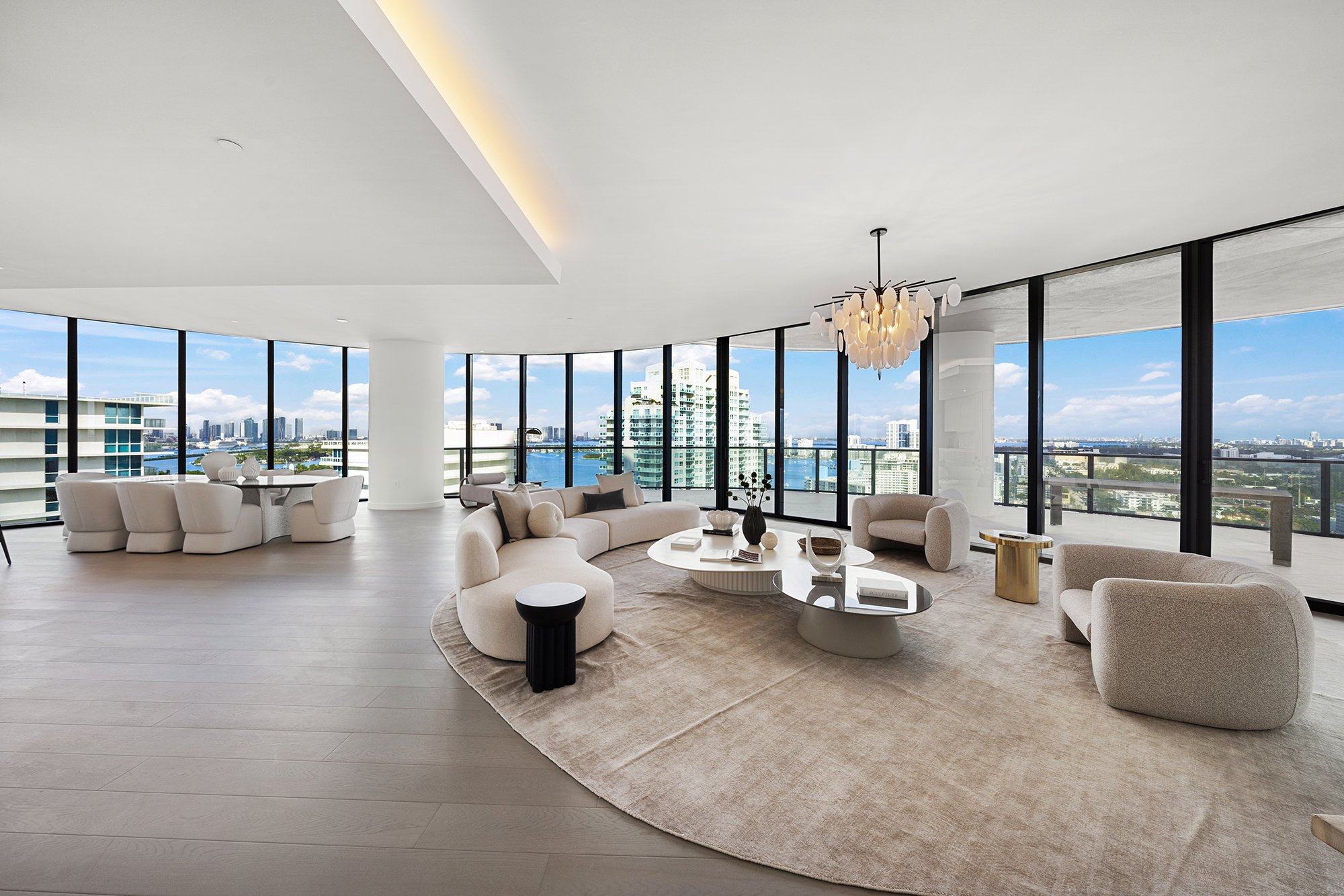 Luxurious modern living room with large windows, cityscape and water views, beige furniture, circular seating, elegant chandelier, and light-colored flooring.