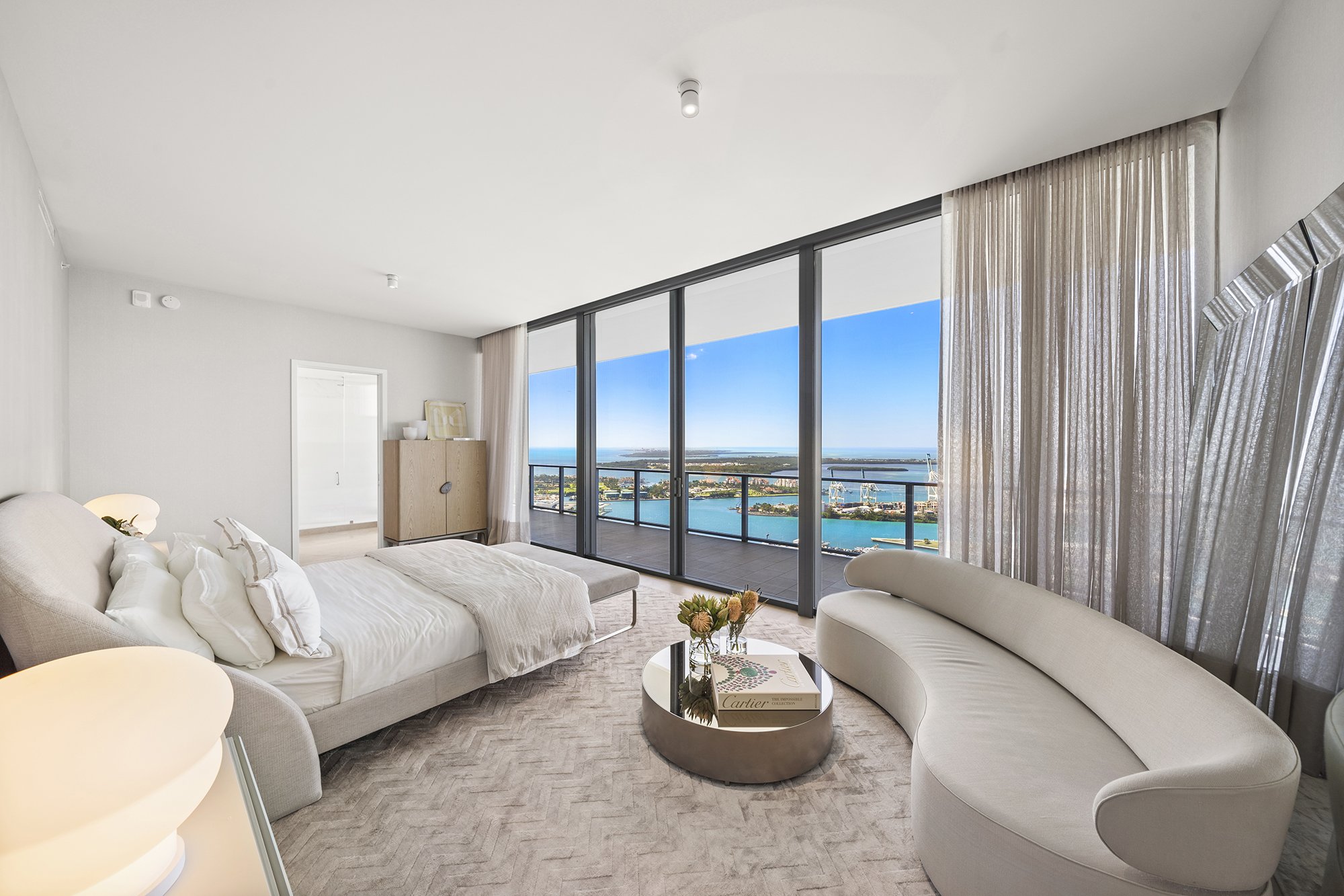 Modern bedroom with large windows offering ocean view, featuring a double bed, curved sofa, round coffee table with books and flowers, neutral color palette, and sheer curtains.