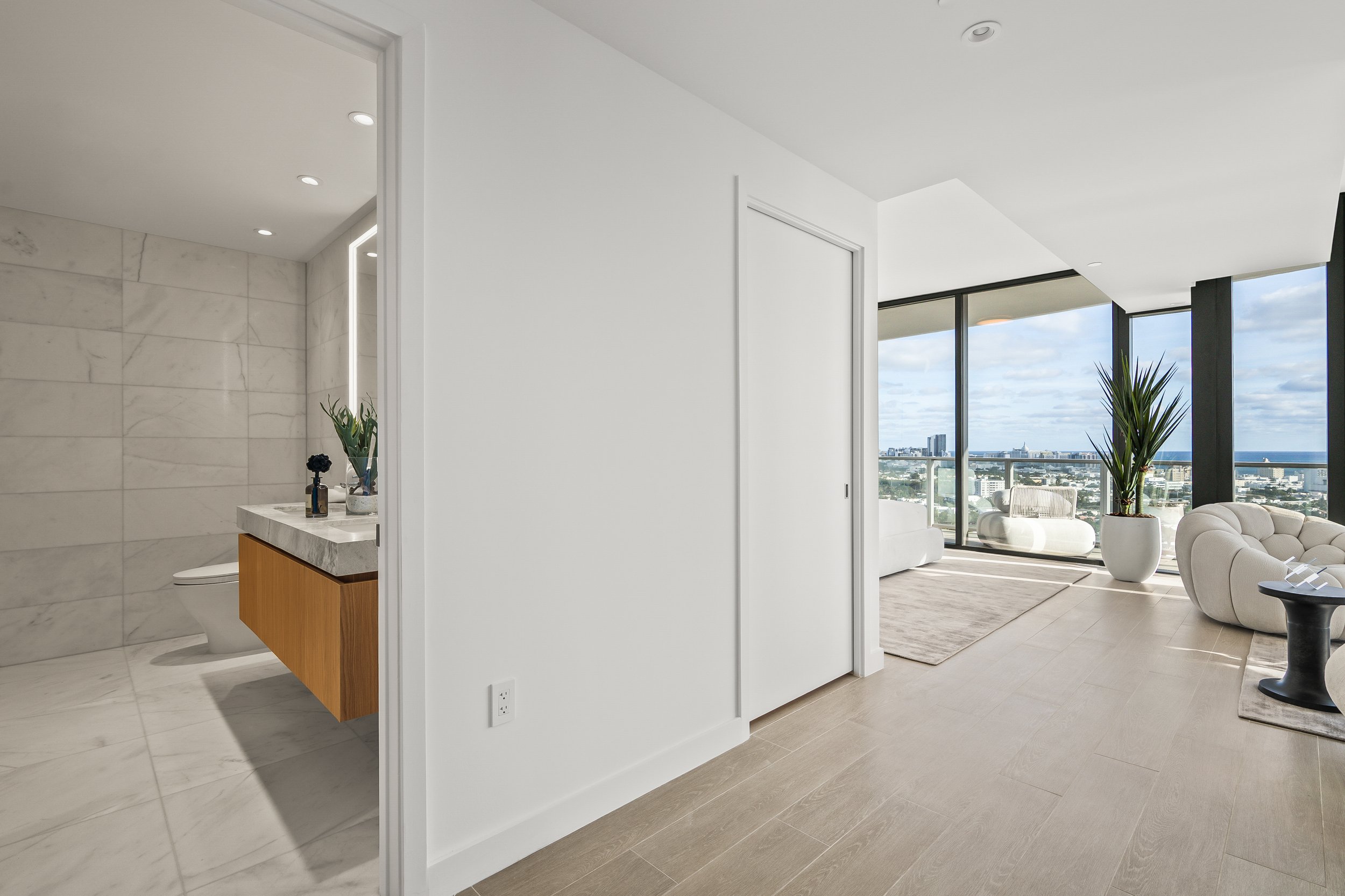 Modern apartment interior with a bathroom and living area. The bathroom has a marble floor and walls with a floating wooden vanity. The living area features large windows with city and ocean views, furnished with white sofas and a potted plant.