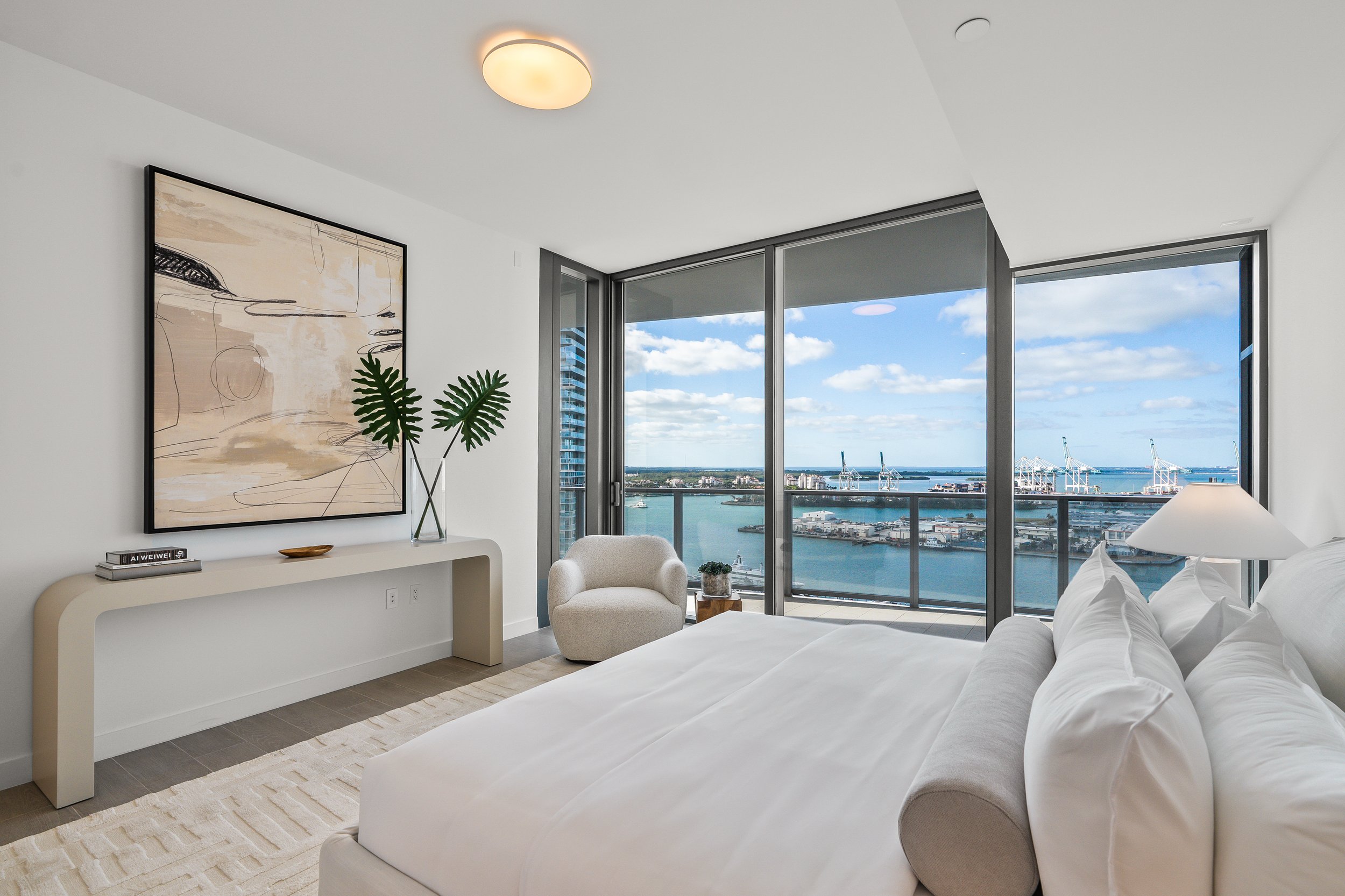 Modern bedroom with large windows offering ocean view, featuring a bed with white linens, a gray chair, a console table with a plant vase, abstract artwork on the wall, and a neutral color scheme.