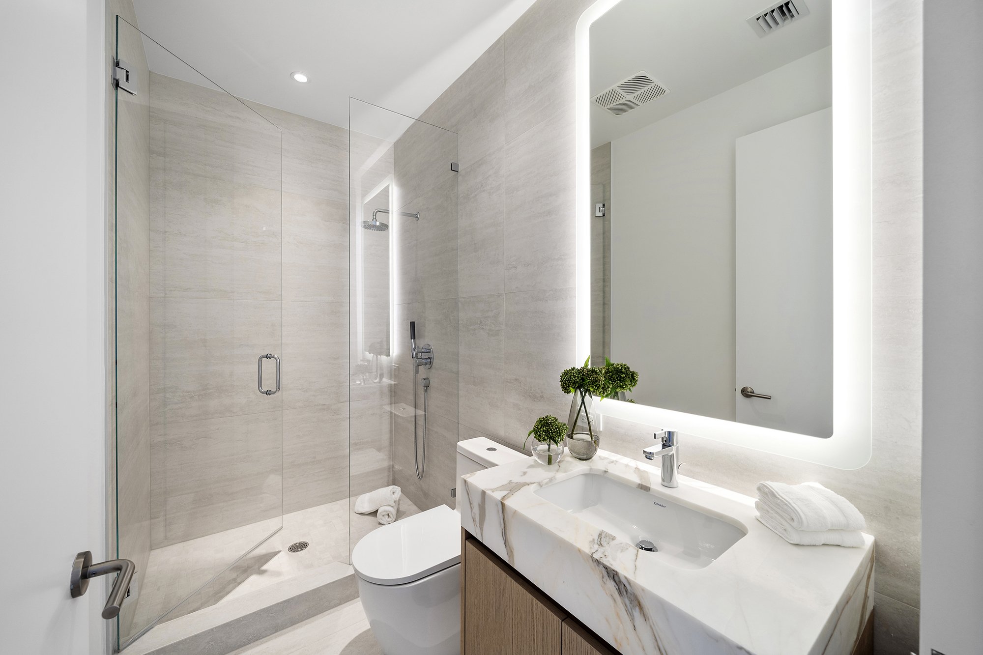 Modern bathroom with glass shower, marble countertop, square sink, backlit mirror, and decorative plants on counter.