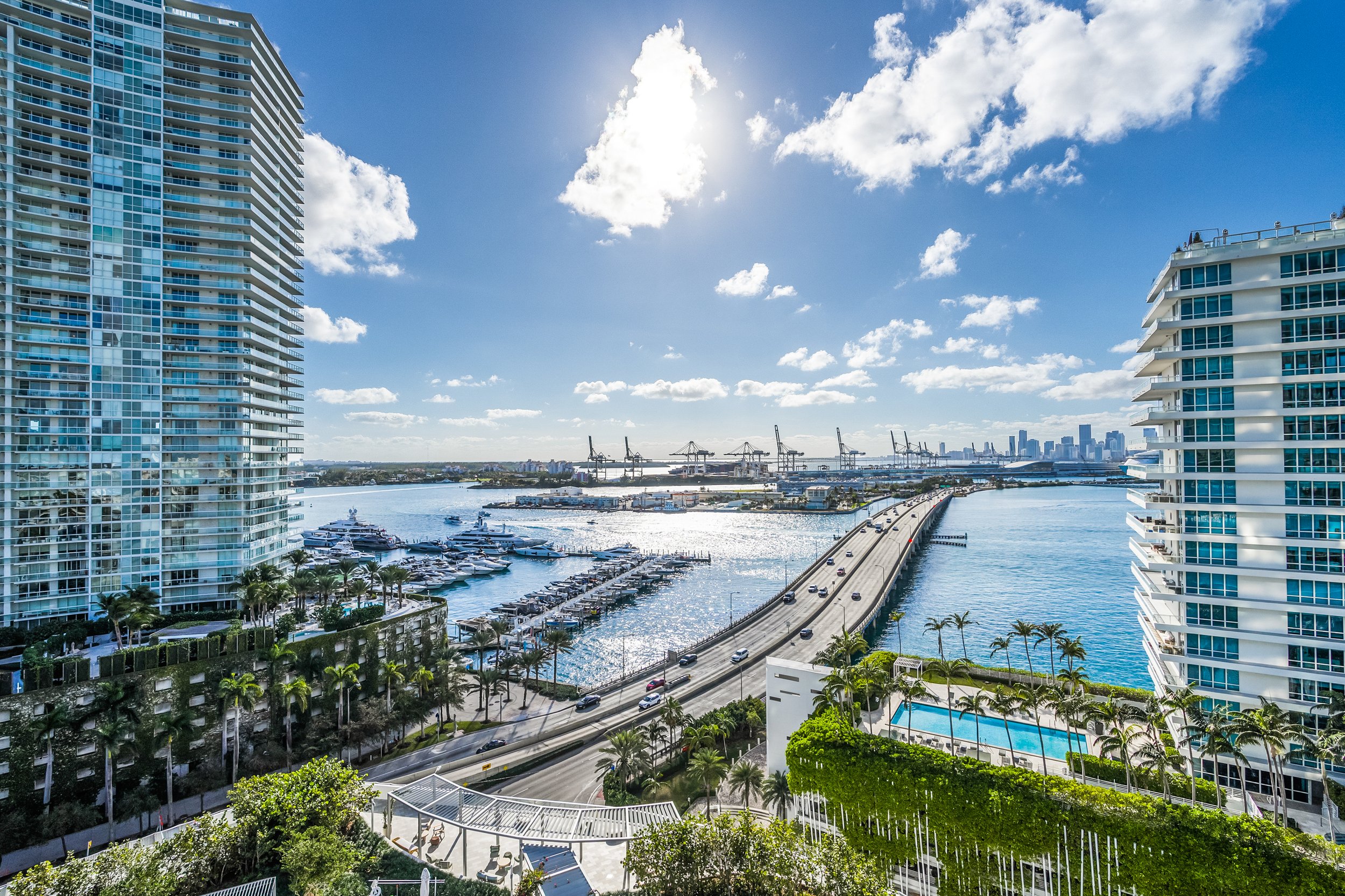 Aerial view of Biscayne Bay with high-rise buildings, marina with yachts, and a bridge connecting to the cityscape of Miami under a bright blue sky.