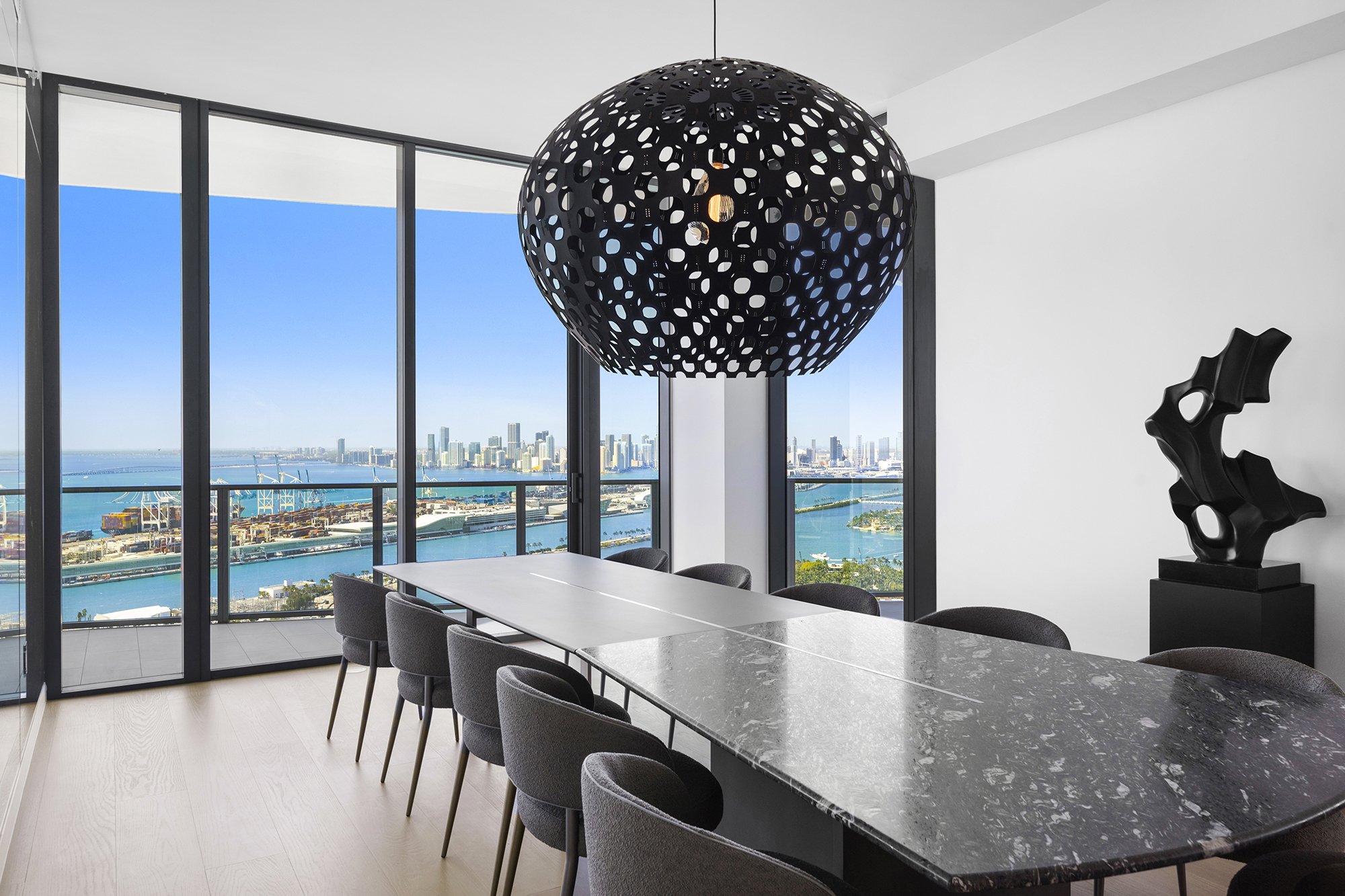 Modern dining room with a large oval marble table, surrounding chairs, a decorative black chandelier, and floor-to-ceiling windows with a city waterfront view.