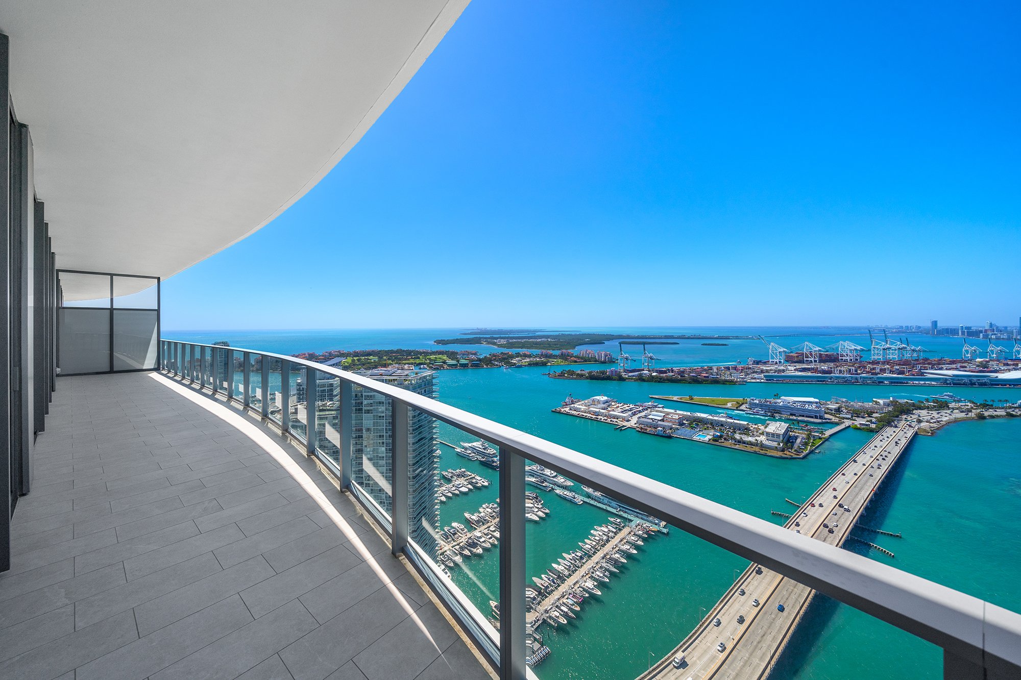 High-rise balcony overlooking city skyline, water, islands, and distant port cranes under a clear blue sky.