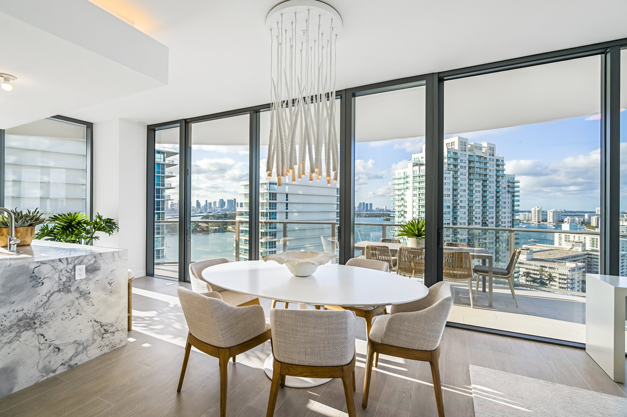 Modern dining area with round table and upholstered chairs, large windows offering city and water views, sleek chandelier, marble kitchen counter, and outdoor seating on the balcony.