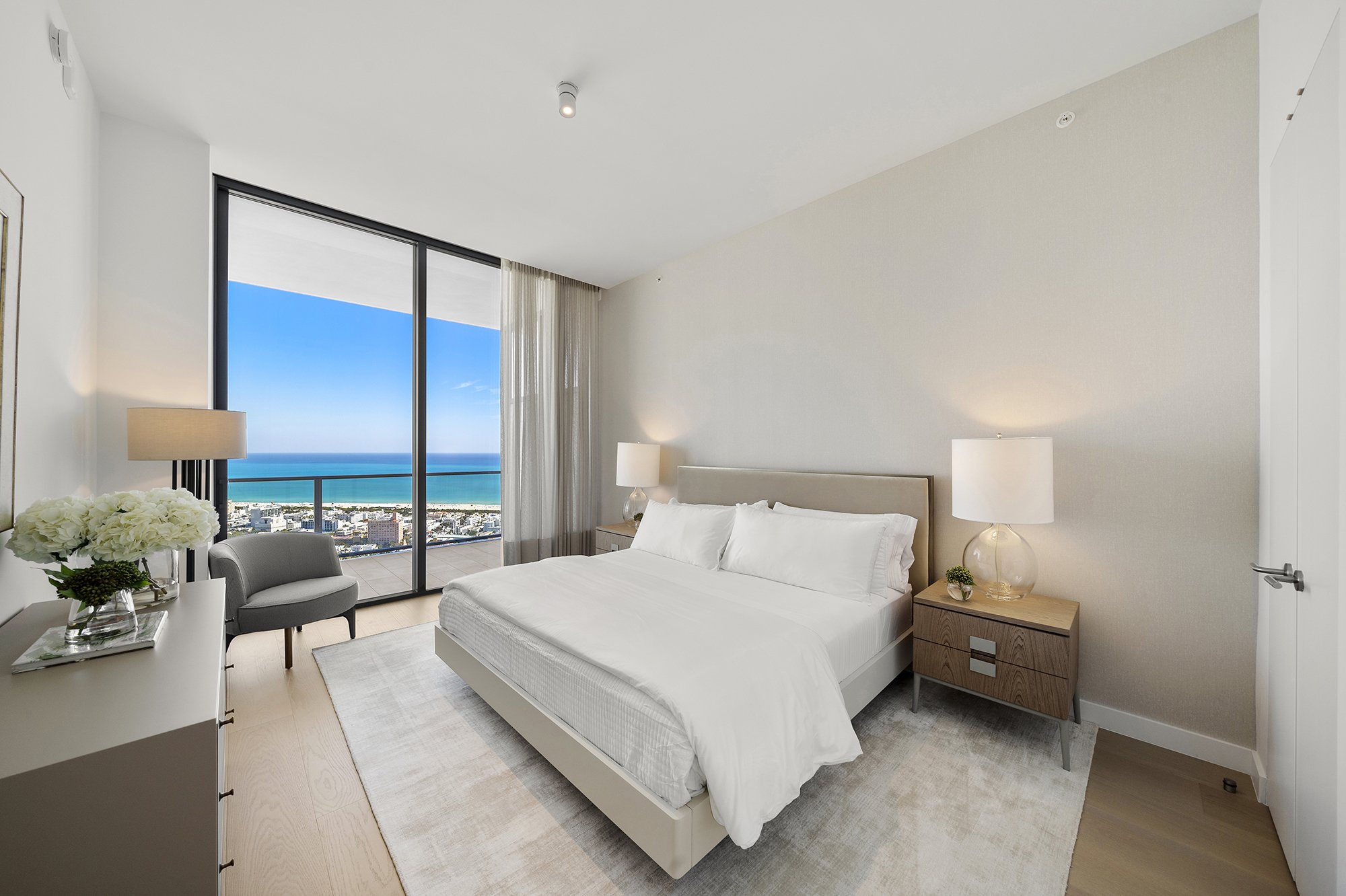 Modern bedroom with ocean view, featuring a large bed, nightstands with lamps, a cozy chair, and a vase of flowers on a dresser, adjacent to floor-to-ceiling window.