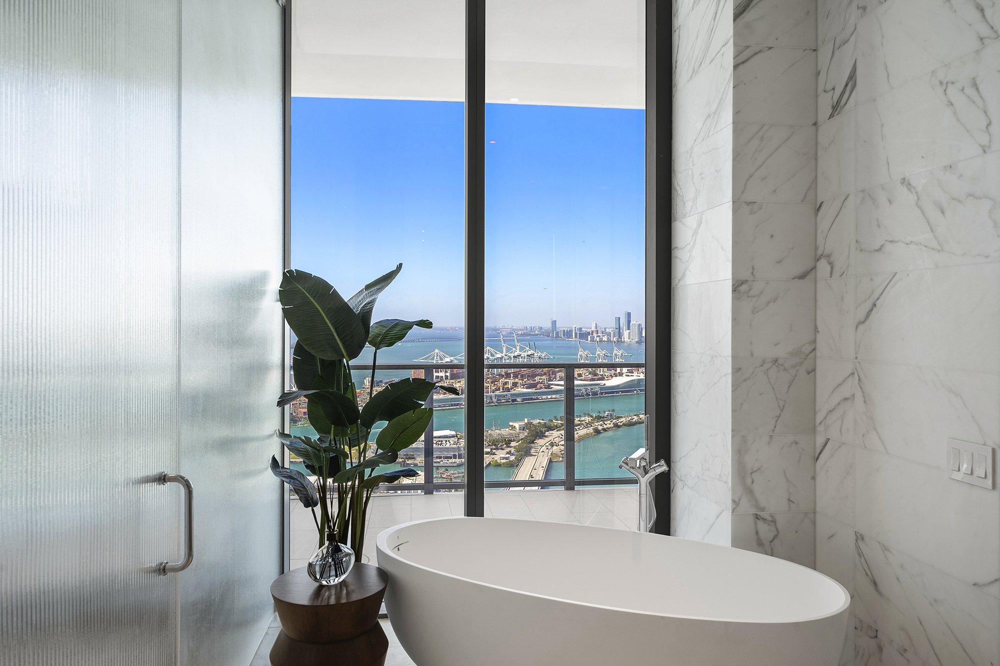 Luxurious bathroom with a freestanding bathtub, marble walls, and a large window offering a city and ocean view, decorated with a potted plant on a wooden stool.