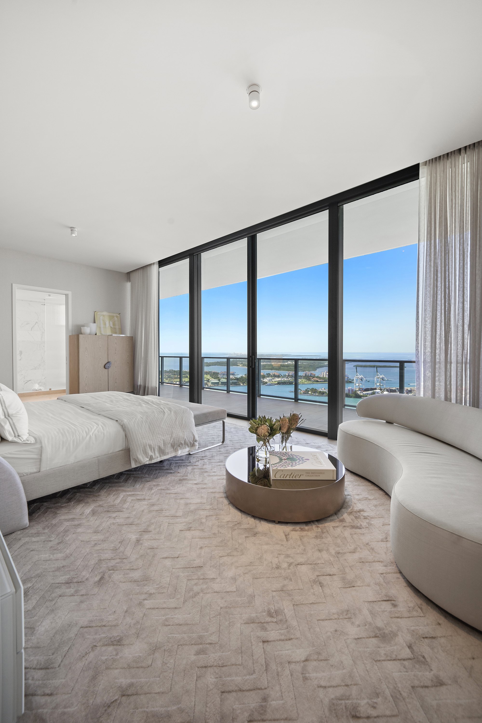 Modern bedroom with large windows offering a view of a cityscape and waterfront, featuring a bed, curved sofa, circular coffee table, and beige carpet.