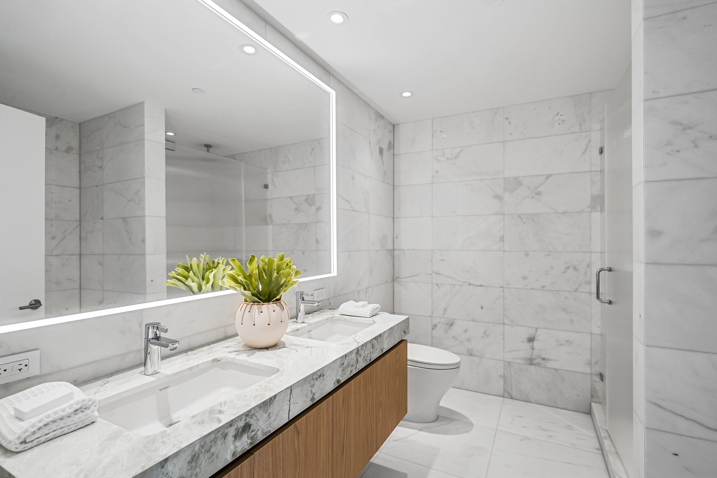Modern bathroom with white marble tiles, dual sinks on a marble countertop, a backlit mirror, a potted plant, towels, and a sleek toilet.