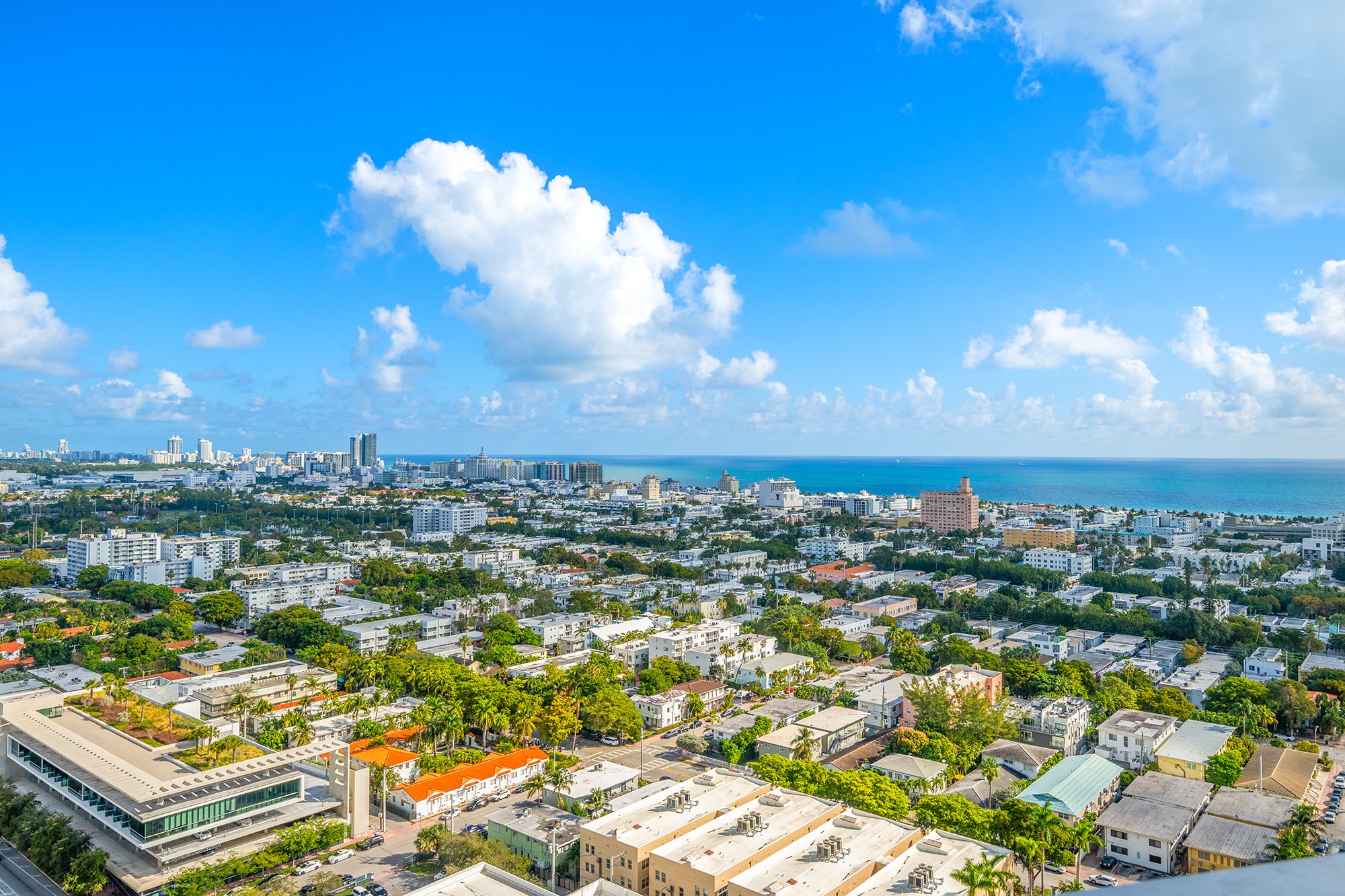 Aerial view of cityscape with ocean in the background, featuring buildings, trees, and a clear blue sky with clouds.
