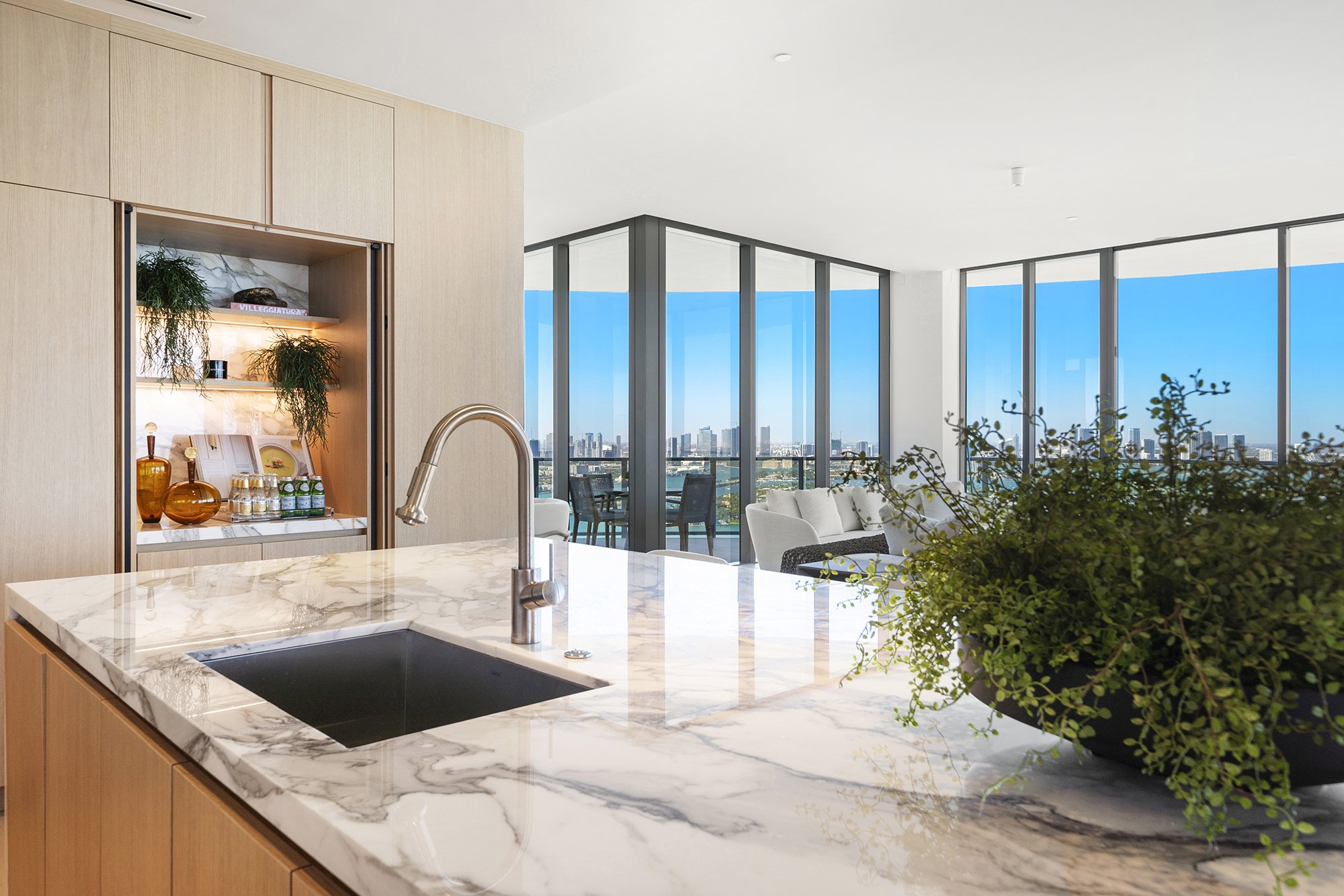 Modern kitchen with marble countertop, open pantry with plants and bottles, overlooking cityscape through large glass windows, with a living area and outdoor balcony.