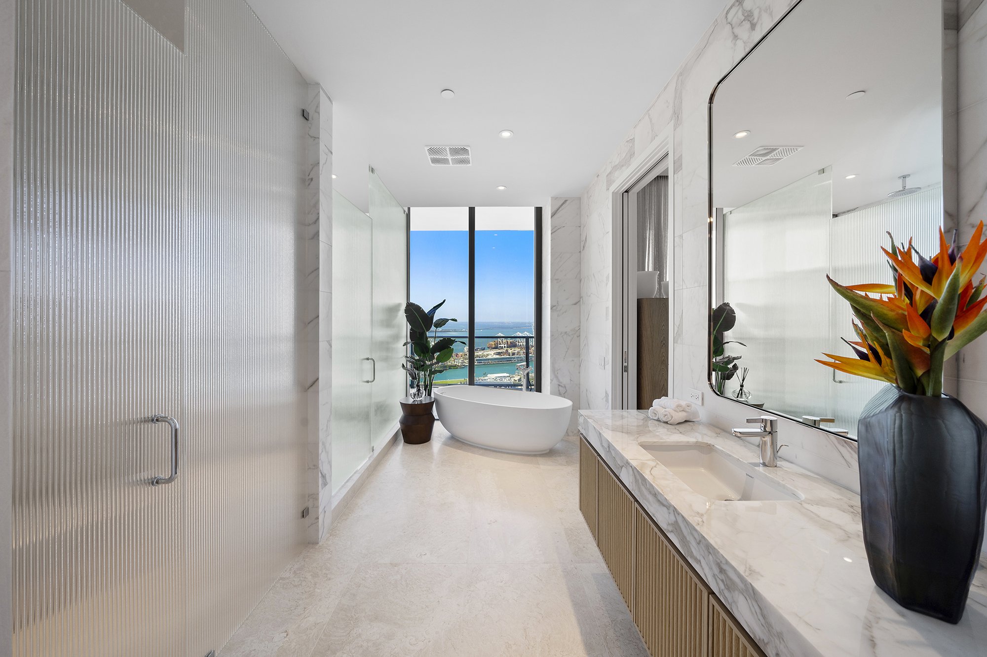 Modern bathroom with a freestanding tub, large mirror, marble counter, potted plant, and glass shower. Ocean view from window.