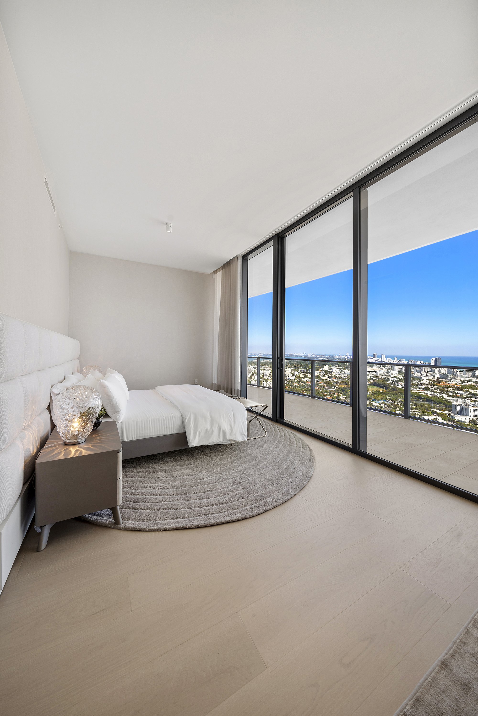 Modern bedroom with minimalist design, featuring a bed with white linens, a nightstand with a decorative lamp, a large circular rug, and floor-to-ceiling windows offering a cityscape view.