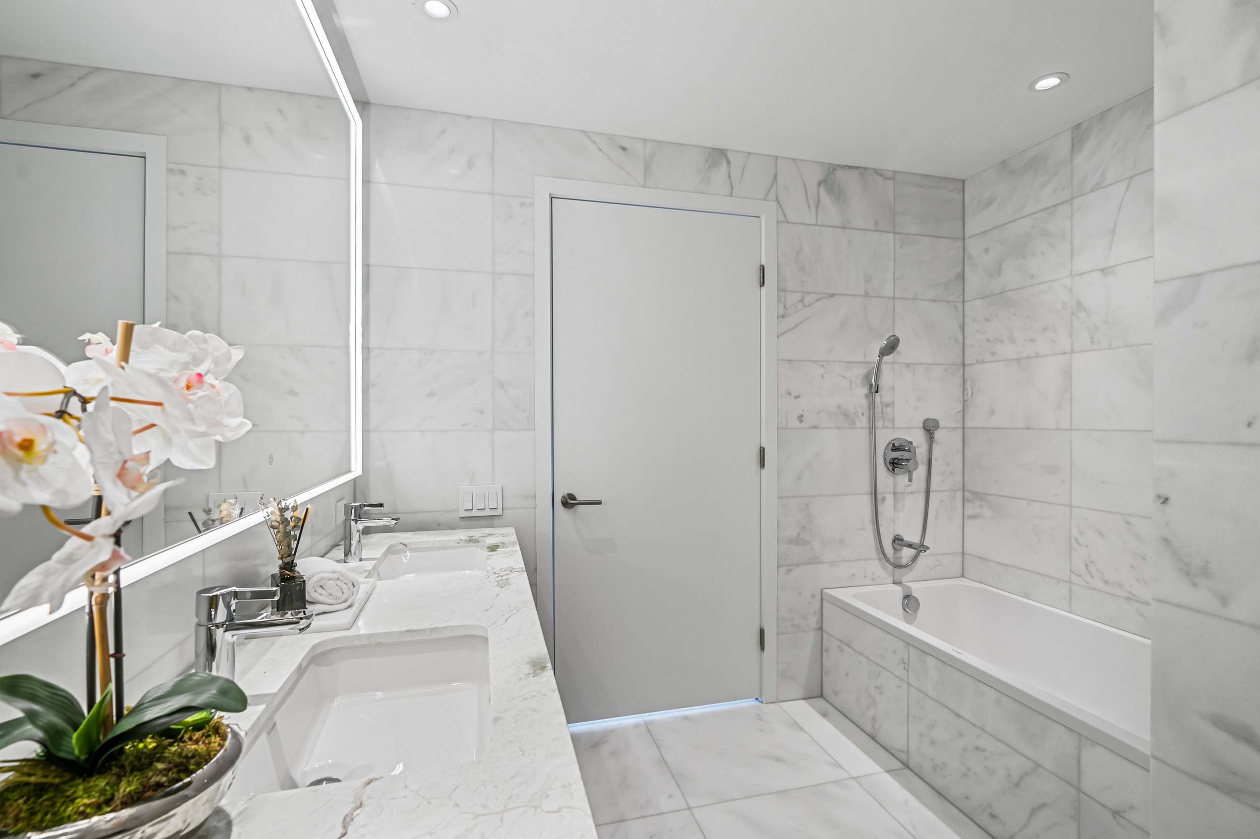 Modern bathroom with marble tiles, double sinks, illuminated mirror, white bathtub, and decorative orchid plant.