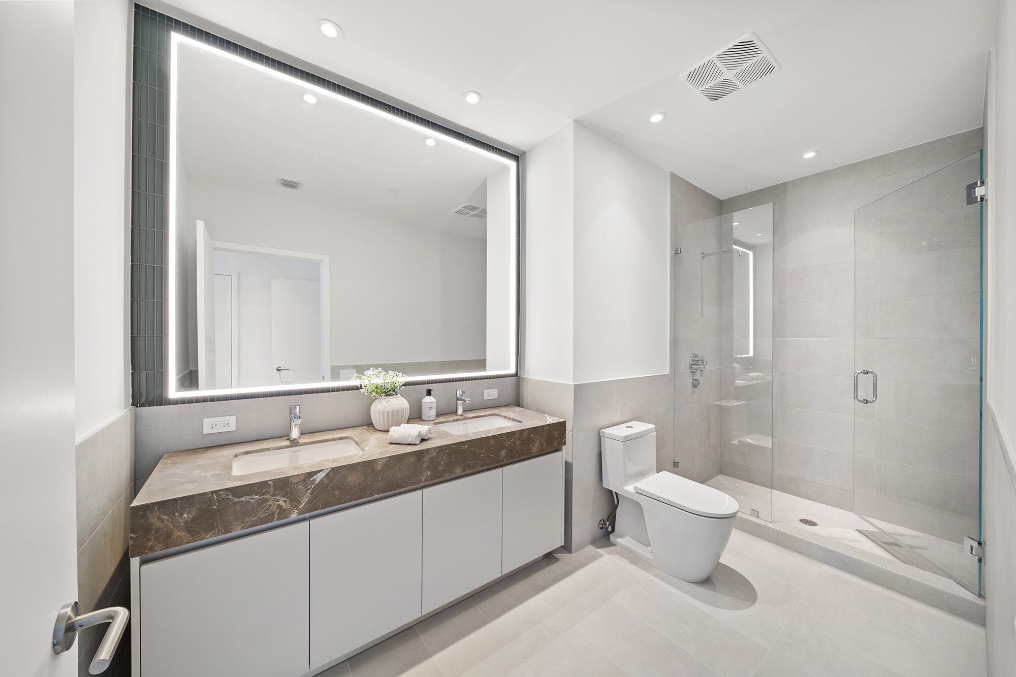 Modern bathroom with large mirror, double sink vanity, toilet, and glass shower enclosure.