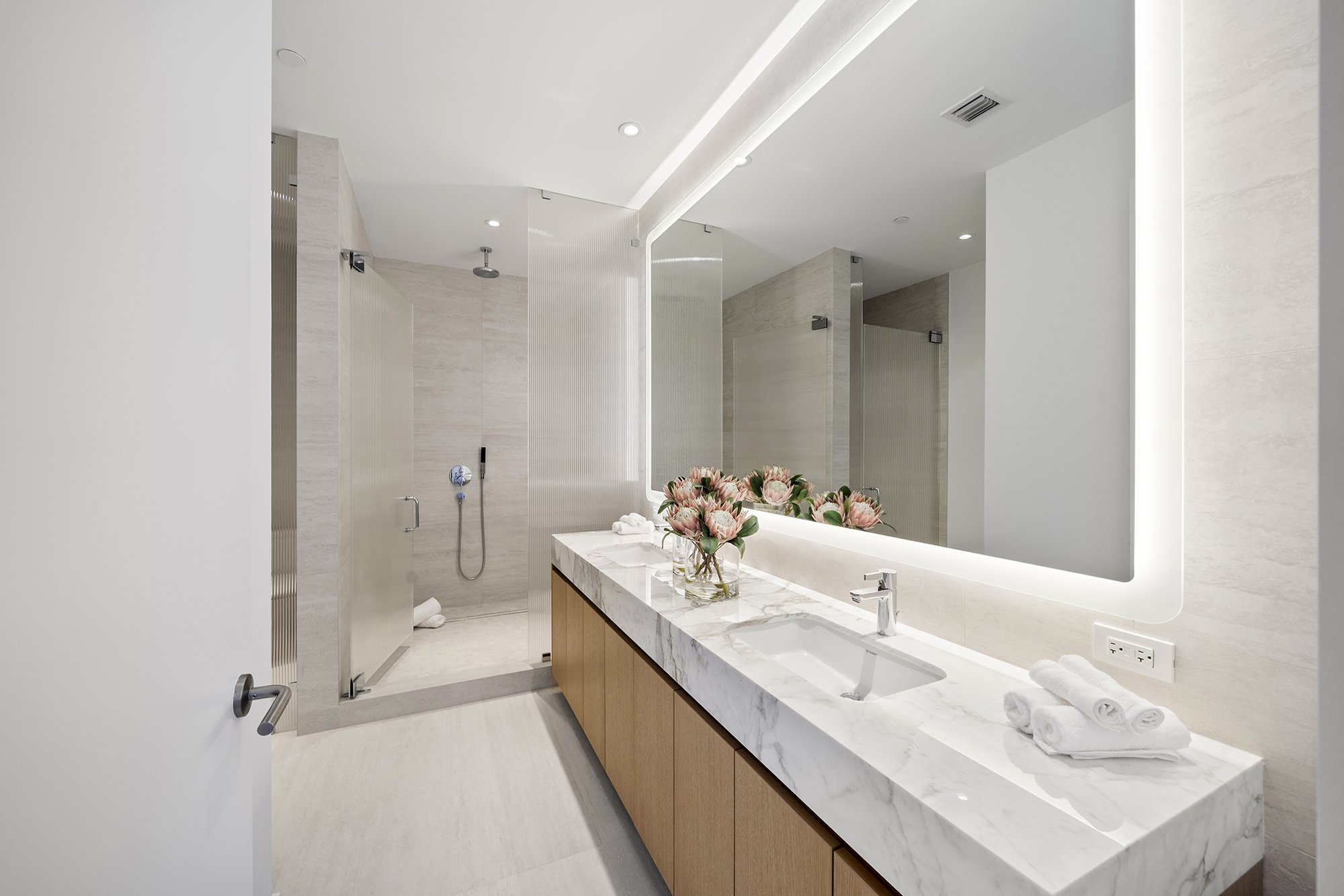 Modern bathroom with a glass-enclosed shower, marble countertop, large backlit mirror, and a vase of decorative flowers.