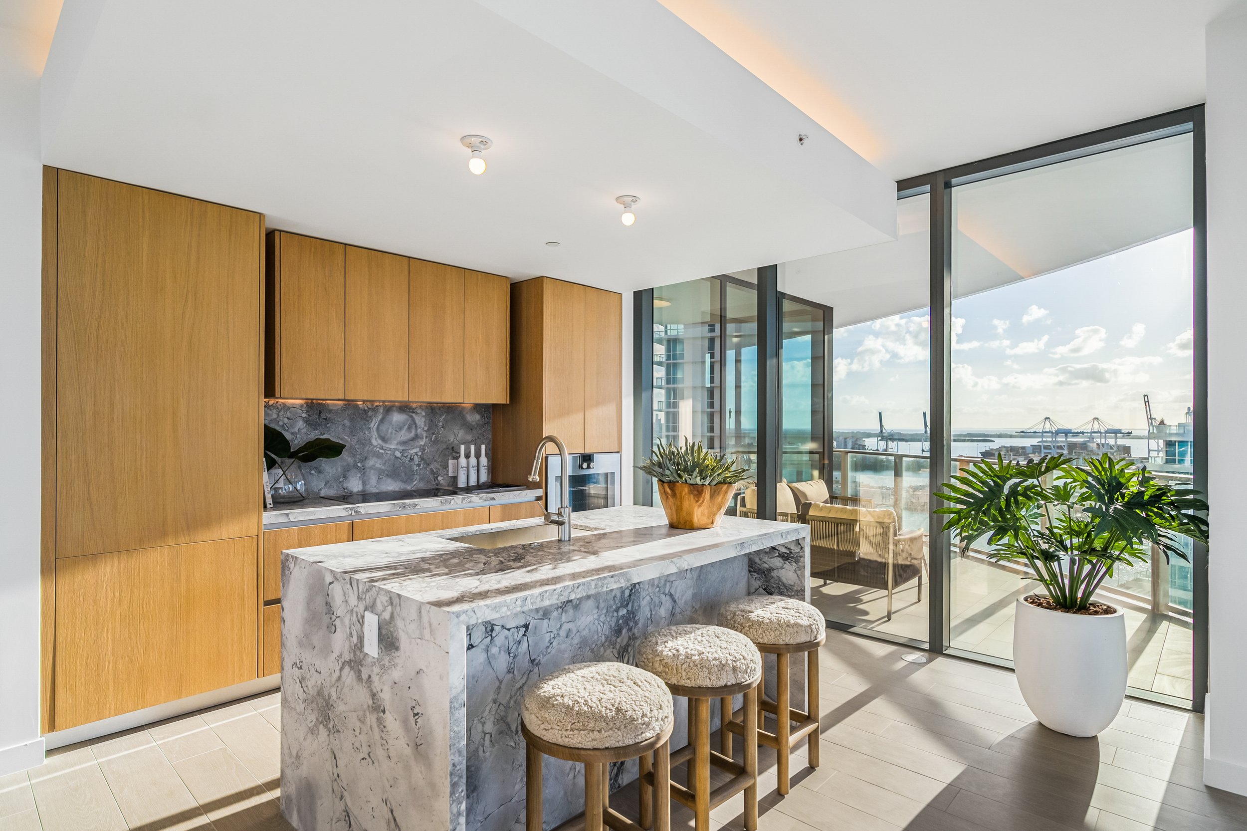 Modern kitchen with wooden cabinets and marble island, featuring stools and a potted plant, adjacent to a glass balcony overlooking a waterfront view.