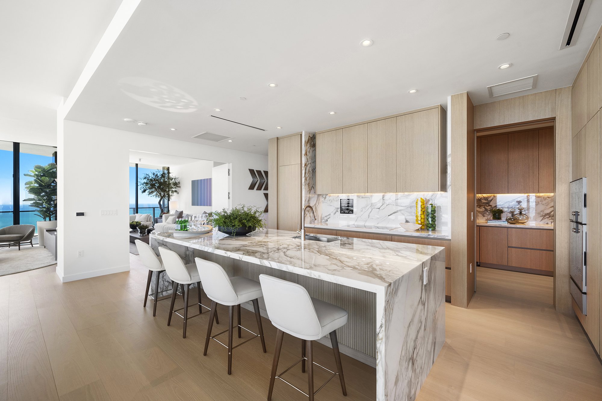 Modern kitchen with marble island, wooden cabinets, and white bar stools, overlooking a living area with large windows and ocean view.
