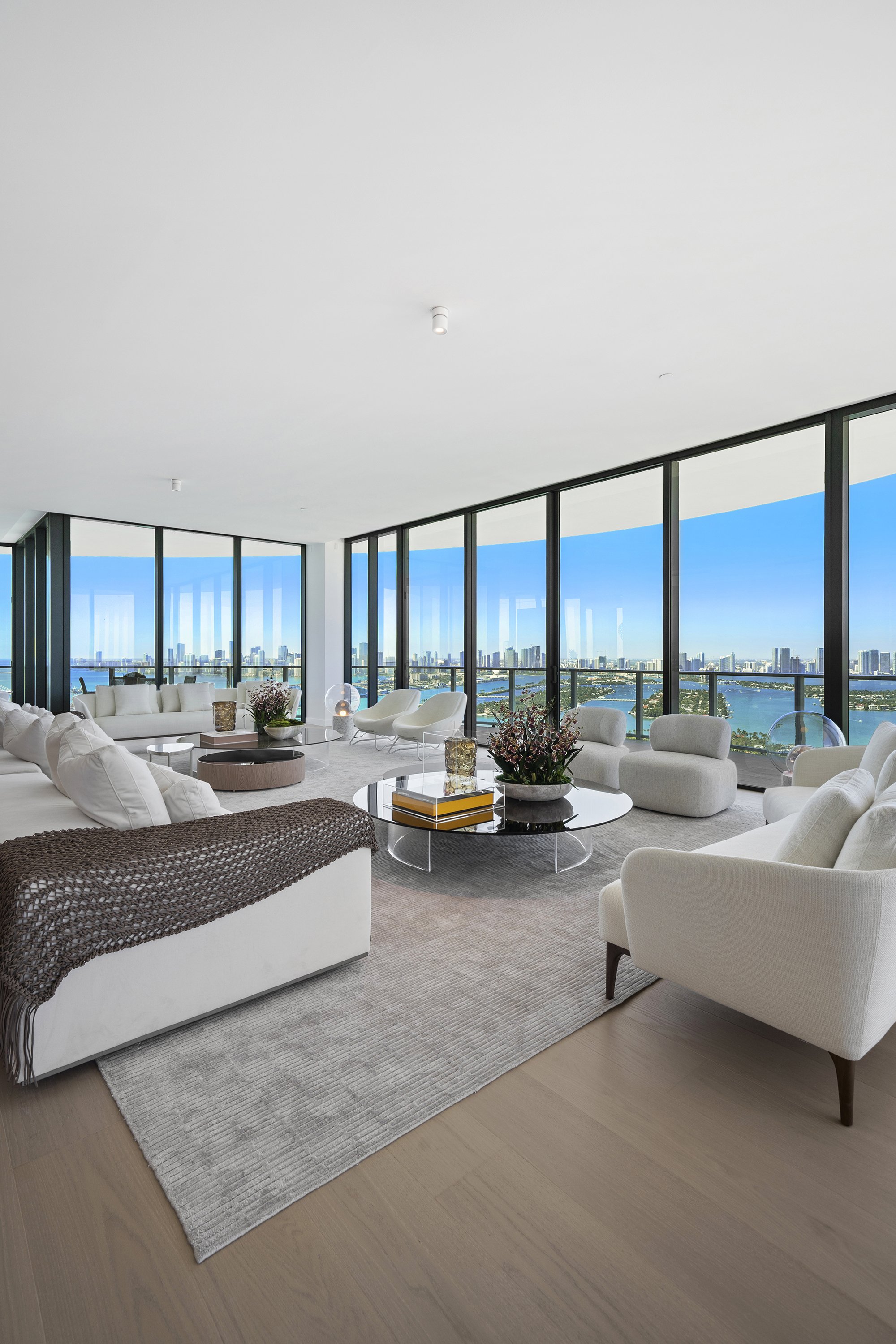 Luxurious living room with modern white furniture, floor-to-ceiling windows, city and water view, large area rug, and contemporary decor on glass coffee tables.