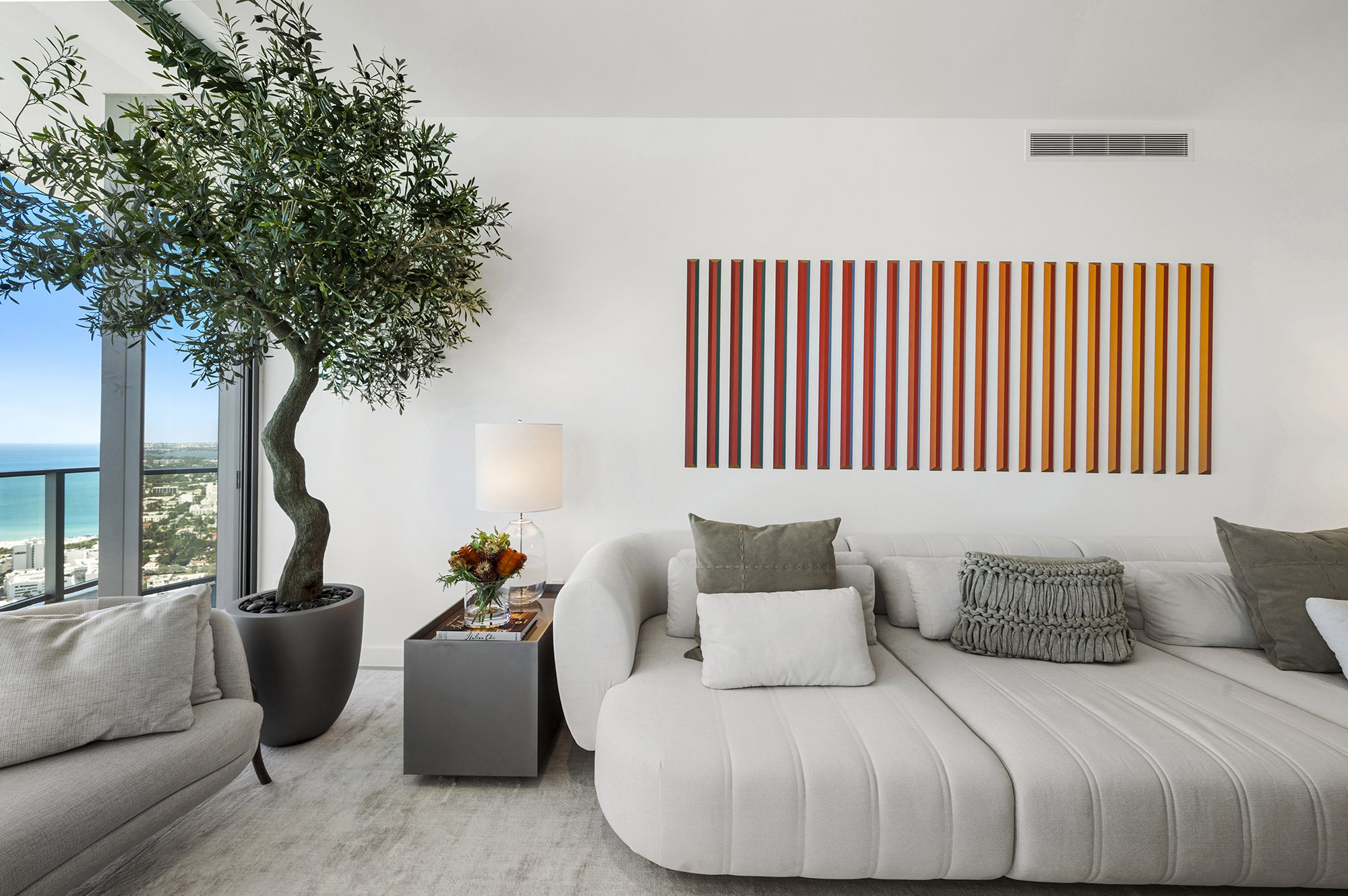 Modern living room with white sofa, decorative pillows, potted plant, colorful striped wall art, and a side table with a lamp and flowers by a window with ocean view.