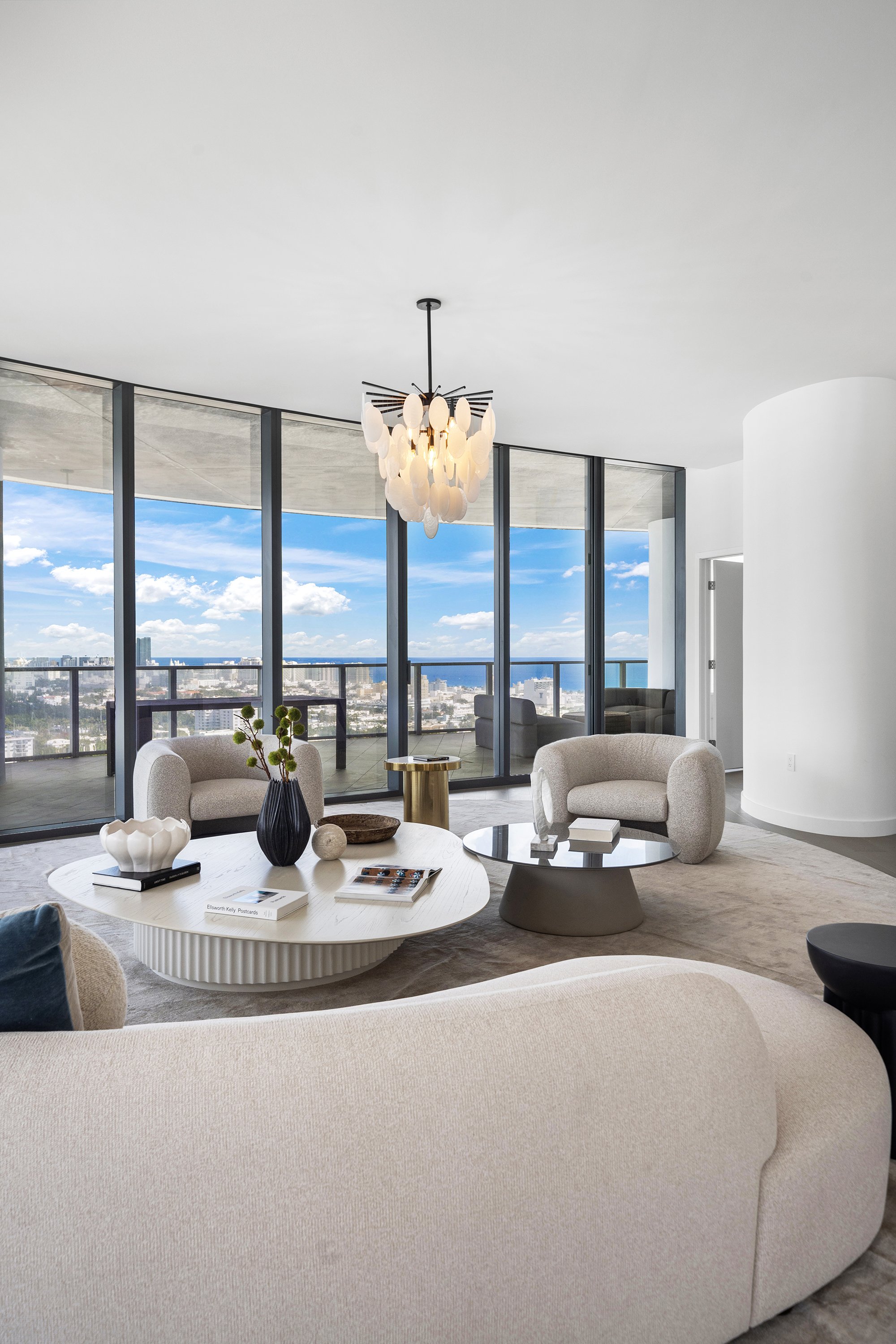 Modern living room with large windows and city view, featuring beige sofas, a round coffee table, decorative items, and a contemporary chandelier.