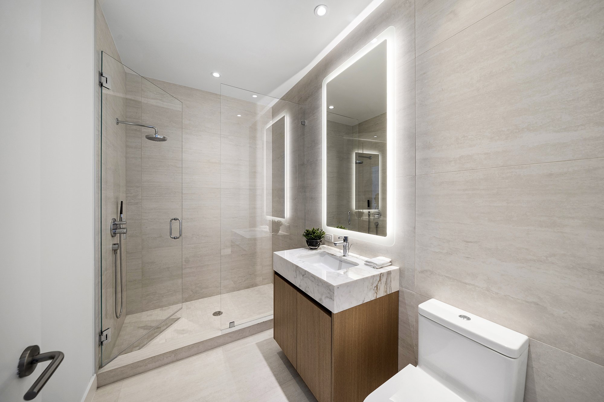 Modern bathroom with a glass shower, lighted mirror, marble sink, and toilet.