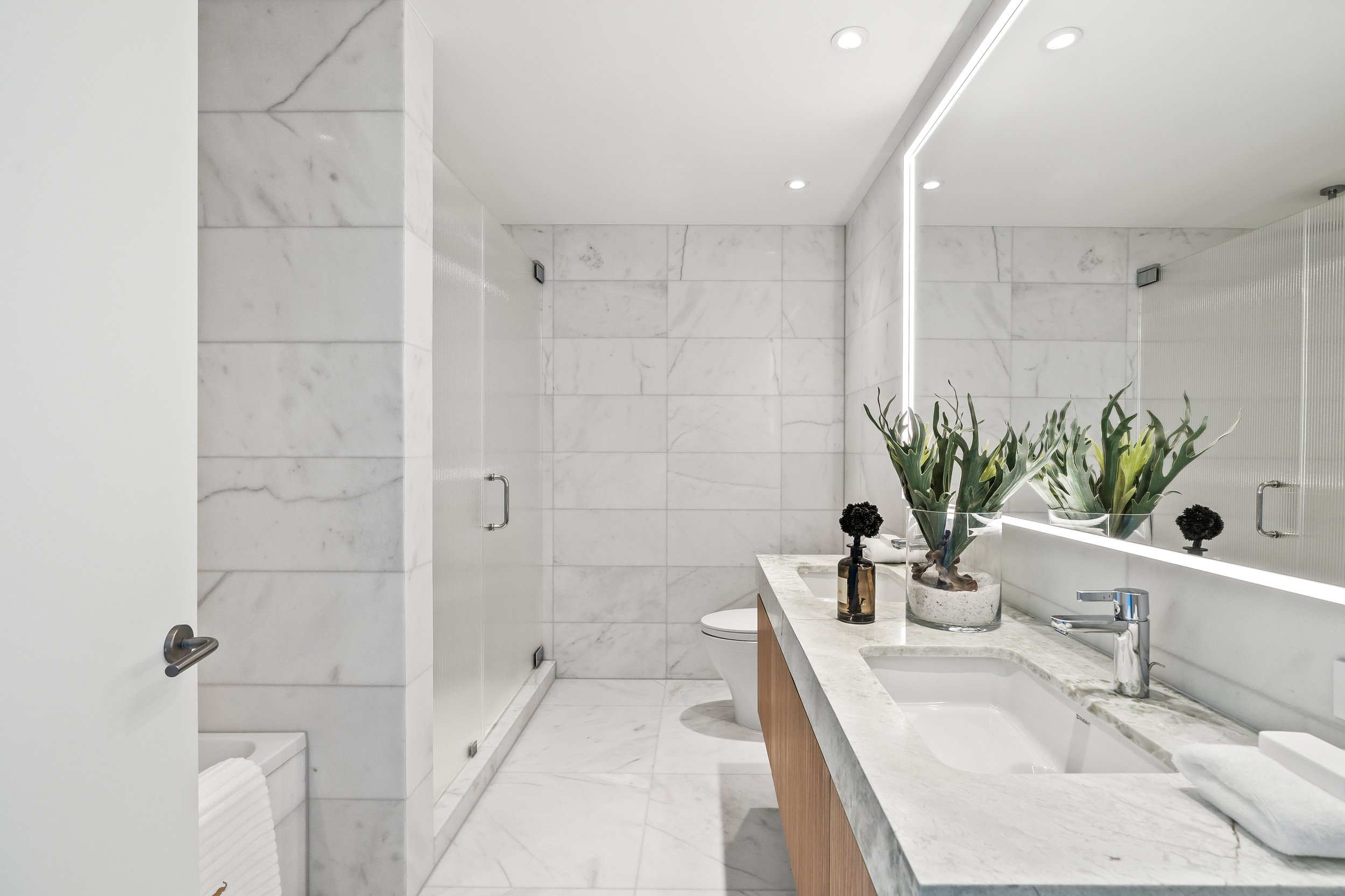 Modern bathroom with marble tiles and countertop, glass shower enclosure, large mirror with integrated lighting, and decorative plants on the vanity.