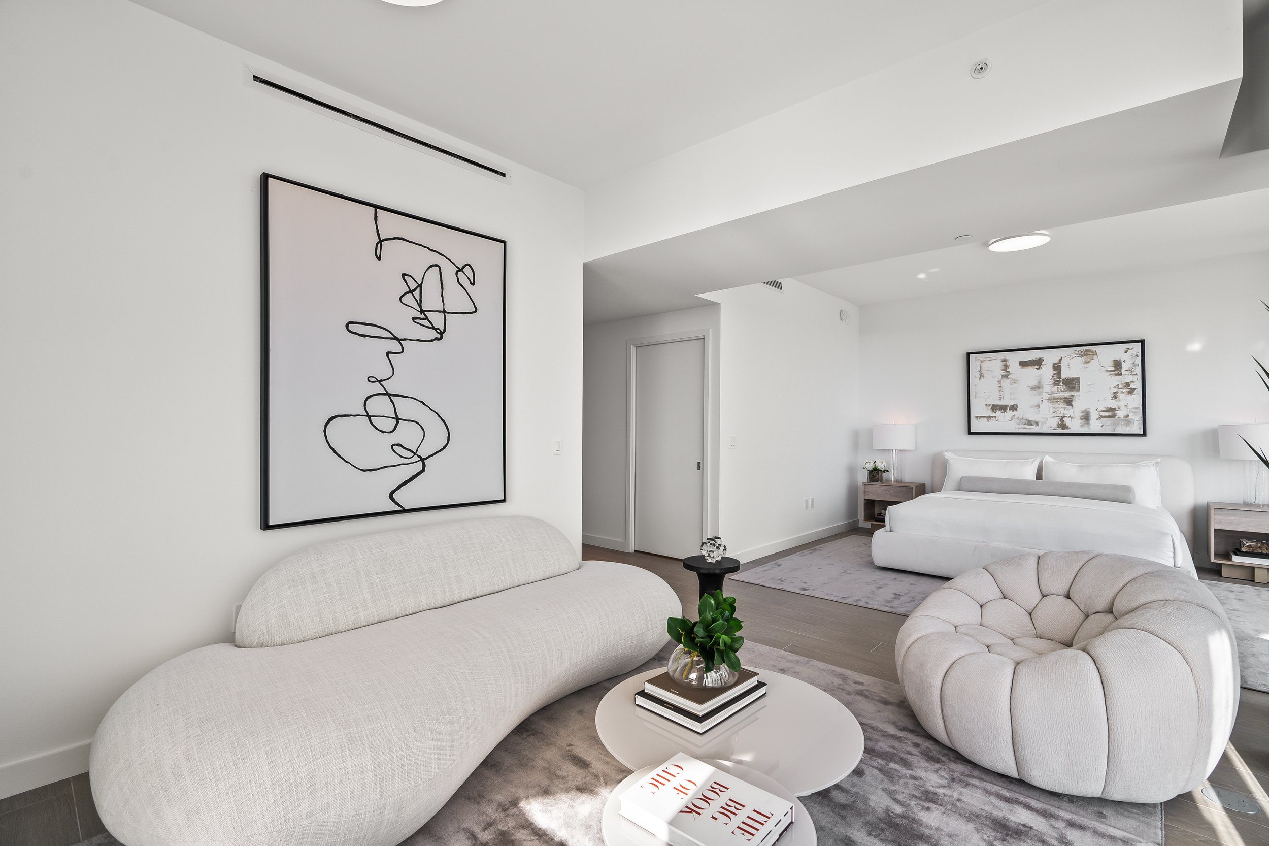 Modern minimalist living room and bedroom with abstract art, a rounded white sofa, armchair, low coffee tables, and a bed with neutral-colored bedding.