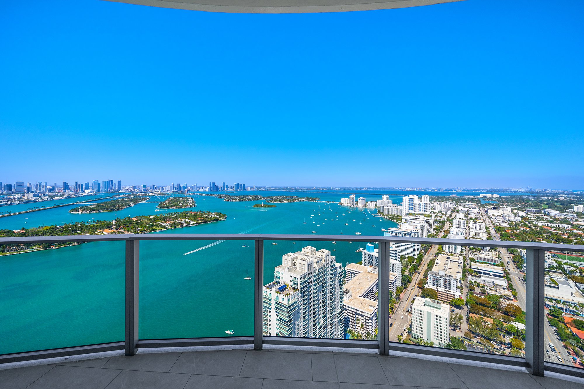 Panoramic view of Miami skyline and Biscayne Bay from a high-rise balcony, with clear blue skies, skyscrapers, islands, and boats on the water.